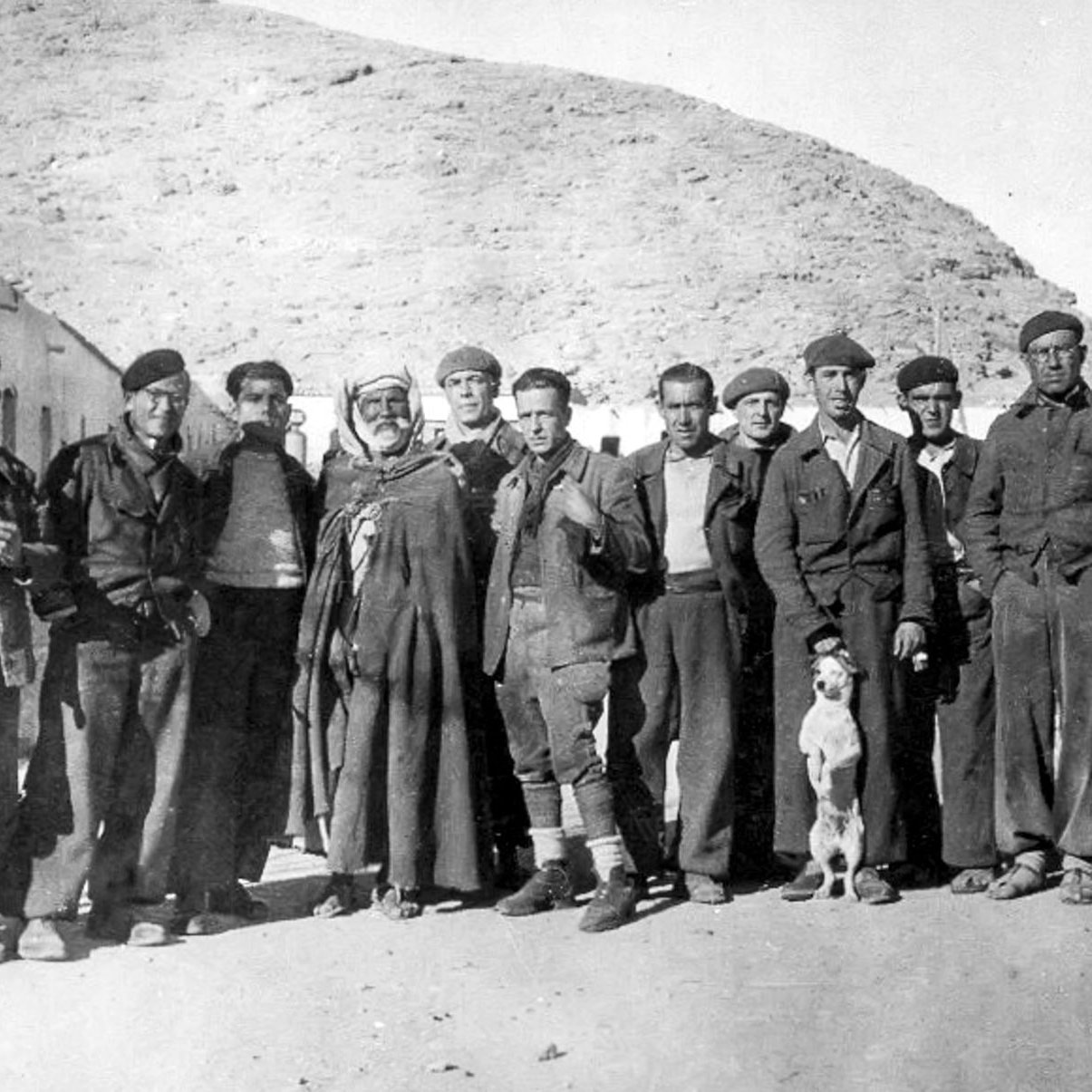 La imagen muestra un grupo de hombres de pie en un entorno rural, posiblemente en una zona montañosa. Están vestidos con ropas tradicionales y de trabajo, algunos con boinas. Hay también un perro que se encuentra entre ellos. La atmósfera es de camaradería, y el paisaje detrás sugiere una época pasada, posiblemente de principios del siglo XX. Los rostros reflejan diferentes expresiones, mientras que la composición transmite una sensación de unión entre los miembros del grupo.
