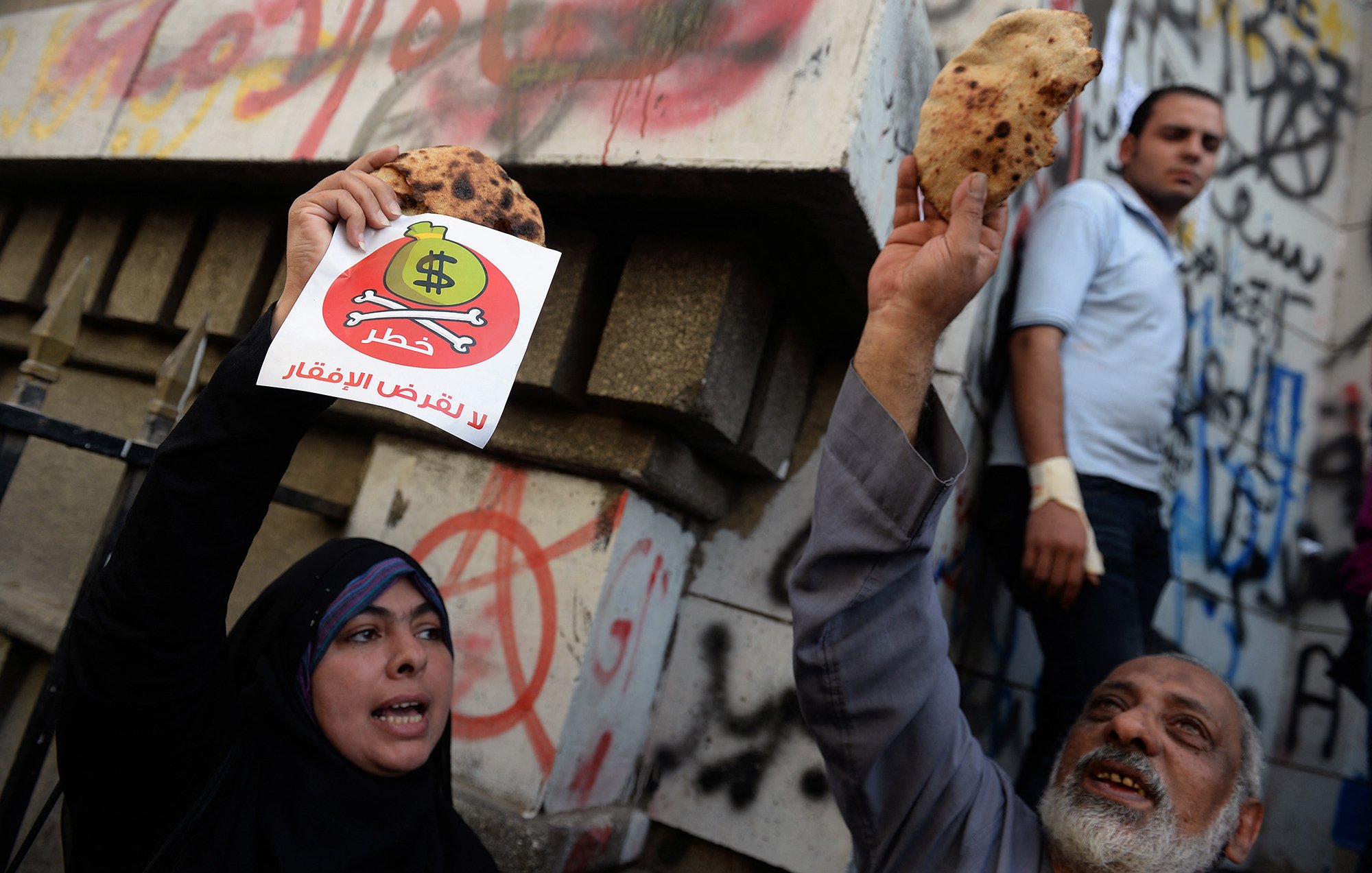 The image depicts a protest scene where individuals are holding up flatbreads and a sign. The sign features a dollar symbol with a prohibition sign over it, conveying a message against inflation or economic hardship, likely related to rising food prices. In the background, there are graffiti-covered walls, indicating a tense atmosphere. The protesters express their discontent, while another person appears in the background observing the scene.
