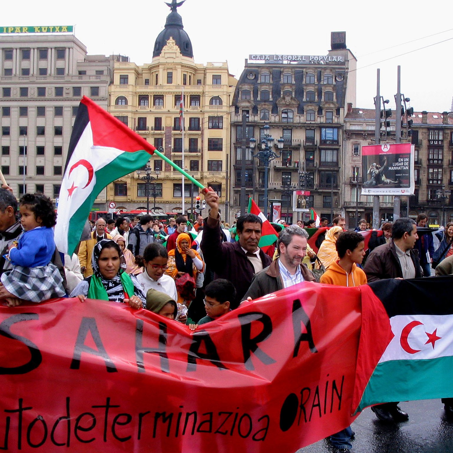 الصورة تُظهر مظاهرة في إحدى الساحات حيث يحمل الناس لافتات وأعلام. يبدو أن المشاركين في المظاهرة يعبرون عن تأييدهم لقضية معينة، حيث تتضمن الأعلام والألوان المستخدمة رموزًا مرتبطة بالصحراء الغربية. المجموعات المختلفة من الناس، بما في ذلك العائلات، يبدون متحمسين وموحدين في مطالبهم. الأجواء تعكس إحساسًا بالوحدة والتضامن.
