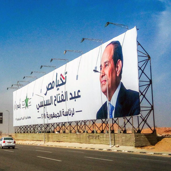 The image depicts a large billboard along a highway featuring a prominent figure, likely a politician, with text in Arabic. The background shows a clear blue sky, and the area appears to be urban, with vehicles on the road. The billboard is designed to attract attention, showcasing both the individual's image and a message or campaign slogan.