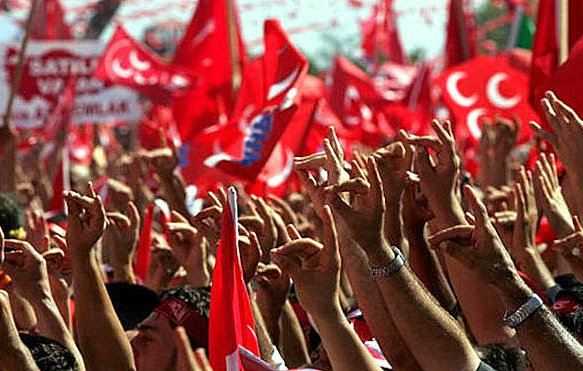 L'image montre une foule rassemblée, brandissant des drapeaux rouges avec des symboles et des motifs typiques. Les participants lèvent les mains, montrant divers gestes de camaraderie ou de soutien. L'ambiance semble festive et engagée, suggérant un événement politique ou une célébration marquée par un fort sentiment nationaliste. Les couleurs dominantes sont le rouge et le blanc, symbolisant souvent l'identité d'un pays.