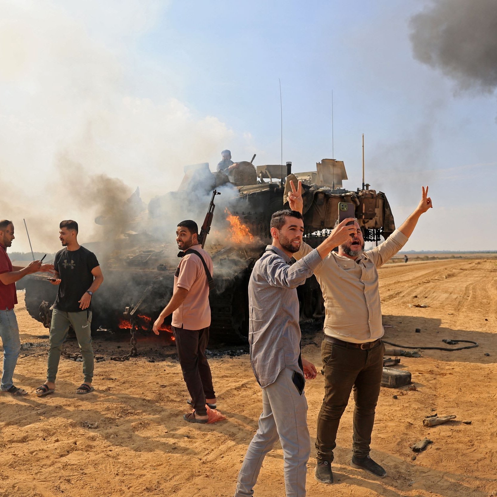 L'image montre un groupe de personnes dans un environnement de conflit. Au premier plan, plusieurs hommes expriment des gestes de victoire ou de protestation, tandis que certains portent des armes. En arrière-plan, on peut voir un véhicule militaire en feu, entouré de fumée noire. Le sol est sablonneux, et le paysage général évoque une situation de tension ou de guerre. L'ambiance est tumultueuse, reflétant un moment de confrontation ou de manifestation.