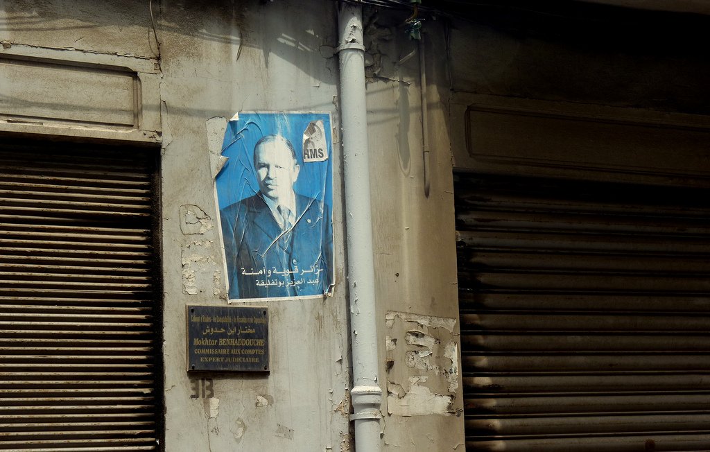 L'image montre un mur d'un bâtiment avec une affiche décolorée d'un homme qui semble être une figure publique. L'affiche est légèrement abîmée, avec des déchirures et des marques. À côté, il y a une plaque signalétique en arabe décorative, elle-même fixée au mur, qui semble indiquer le nom d'un établissement ou d'une personne liée au bâtiment. Le mur présente également des signes d'usure, avec une peinture écaillée et quelques éclats de surface. Les volets en métal, qui sont fermés, ajoutent à l'atmosphère urbaine de la scène.