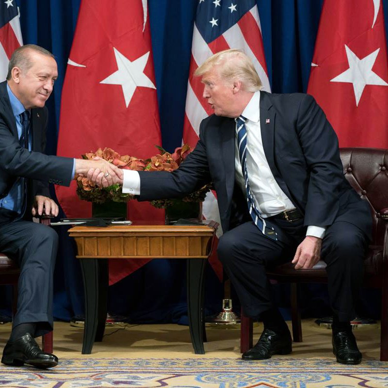 تظهر الصورة لقاءً رسميًا بين شخصيتين بارزتين، حيث يجلس كل منهما في كرسي ويقومون بمصافحة بعضهم البعض. الخلفية تحتوي على علمين، أحدهما يمثل الولايات المتحدة والآخر يمثل تركيا. يظهر في الصورة وضع تعاوني، مما يدل على أهمية اللقاء بين الطرفين. يمكن رؤية أيضًا بعض الزهور على الطاولة بينهما، مما يضيف لمسة من الأناقة إلى المشهد.