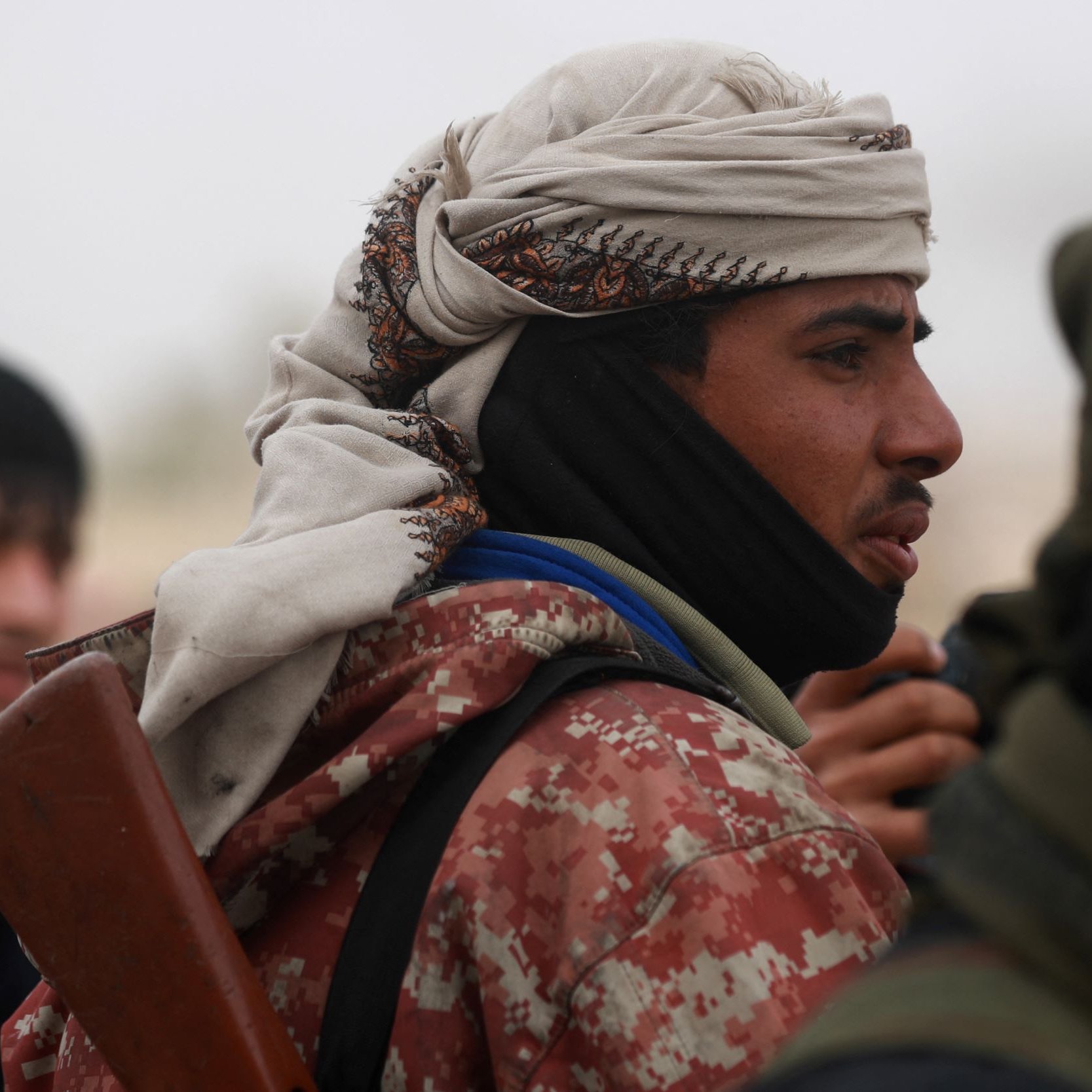 در این تصویر، یک فرد در حالتی نزدیک به دوربین قرار دارد. او کلاهی بر سر دارد و چهره‌اش جزئیاتی را نشان می‌دهد که به او ظاهری خاص می‌دهد. لباسش نیز شامل یک روپوش است که الگوهای خاصی دارد. در پس‌زمینه، افرادی دیگر نیز دیده می‌شوند. جو تصویر احتمالاً جدی و نظامی است.