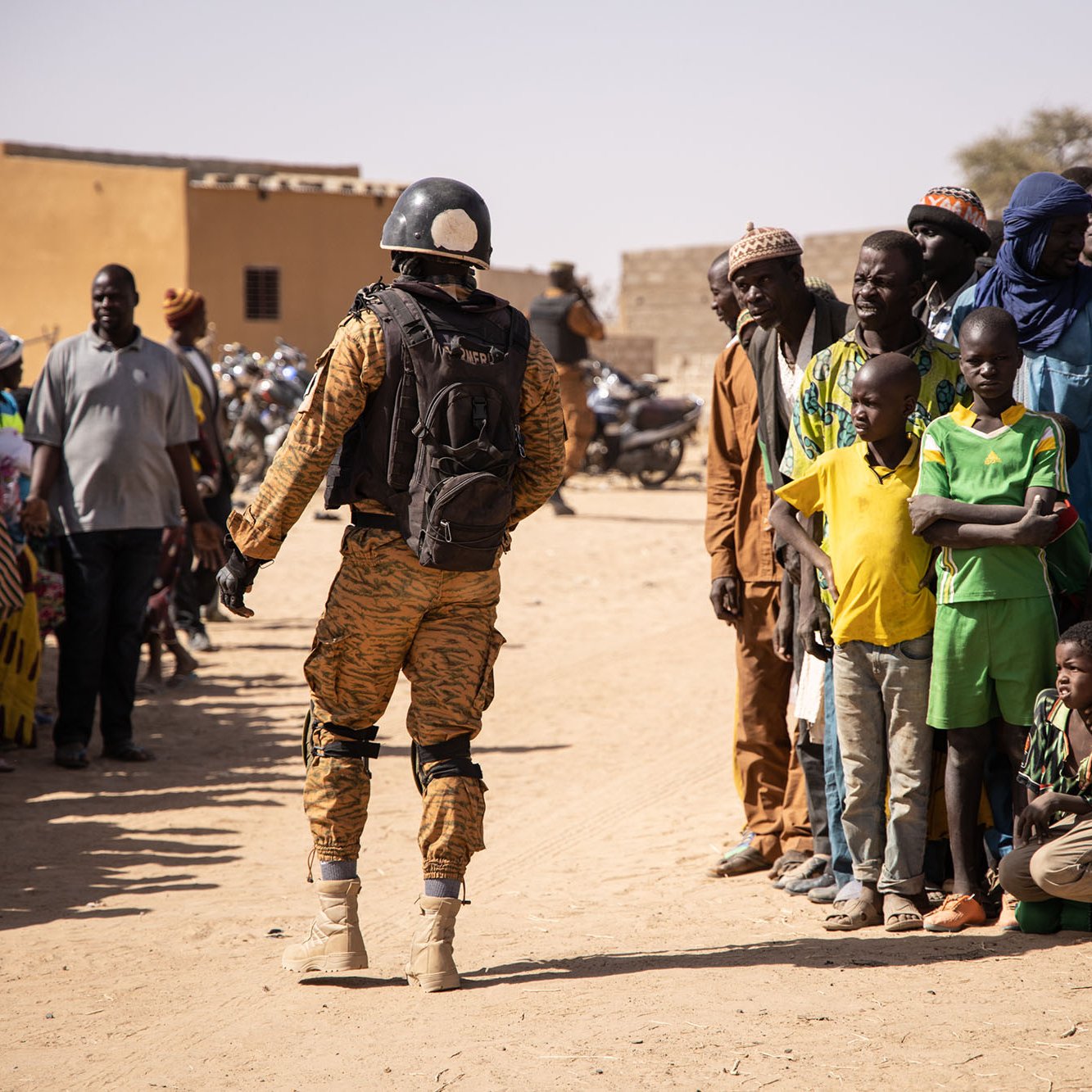 L'image montre un soldat en uniforme militaire, portant un casque et un sac à dos, se tenant face à une foule. Les personnes dans la foule sont de différentes tranches d'âge, allant des enfants aux adultes, et semblent observer le soldat avec attention. Le décor évoque une ambiance de village rural, avec des bâtiments simples et un sol en terre battue. Les couleurs de l'uniforme du soldat contrastent avec celles des vêtements des membres de la foule. L'atmosphère générale semble être celle d'une interaction, peut-être dans le cadre d'une mission de sécurité ou d'assistance.