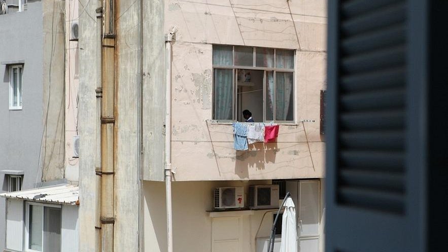 L'image montre un immeuble avec des murs de couleur rose. À un des balcons, on aperçoit un enfant qui semble regarder à l'extérieur. Des vêtements sont étendus sur la rambarde du balcon, et au sol, on peut voir un parasol ouvert. À droite, un volet est partiellement ouvert, ajoutant une autre dimension à la scène. L'ambiance semble calme et typique d'un environnement urbain.