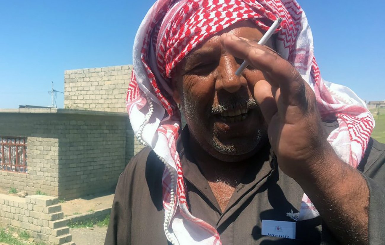The image shows a man wearing a traditional headscarf, often referred to as a keffiyeh or ghutrah, with a red and white pattern. He is standing outdoors, possibly in a rural area, with stone structures visible in the background. The man appears to be smiling or in a candid moment, holding a cigarette between his fingers. The setting is bright, indicating a sunny day, and the landscape around him includes some greenery and dirt paths.