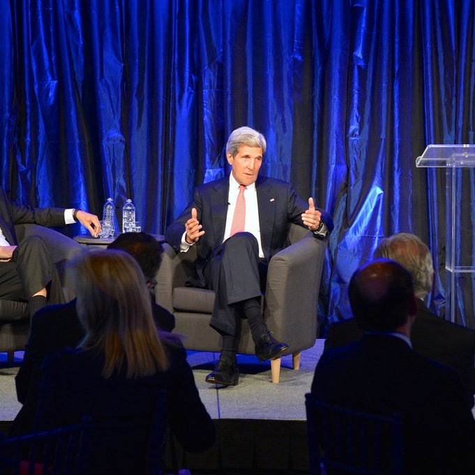 L'image montre une scène de conférence ou de discussion, où deux intervenants sont présents sur une scène. À gauche, un homme est assis sur un fauteuil, écoutant attentivement, tandis qu'à droite, un autre homme parle avec des gestes, probablement en train de partager ses idées ou répondre à des questions. La scène est éclairée avec un fond de rideaux bleus, créant une ambiance professionnelle et formelle. Il y a également un podium avec un microphone, prêt à être utilisé. En bas, on peut apercevoir un public assis, attentif à la conversation.