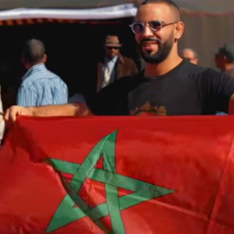 En la imagen se puede ver a un grupo de personas en un ambiente al aire libre, posiblemente en un evento o celebración. Un hombre sostiene una bandera de Marruecos, que es de color rojo con una estrella verde en el centro. Otros hombres también están presentes en el fondo, algunos sosteniendo banderas o participando en la actividad. La escena refleja un ambiente de camaradería y orgullo nacional.