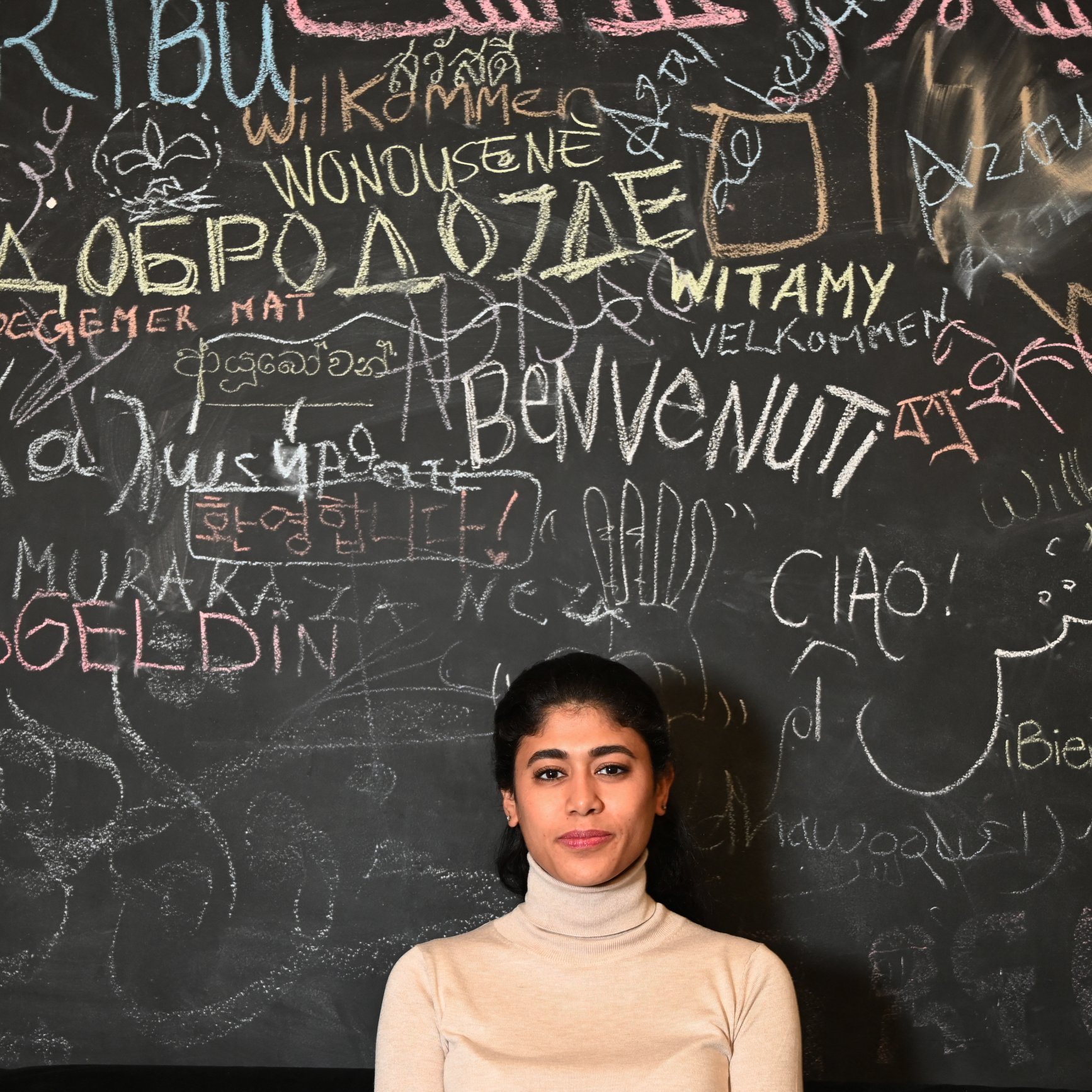 L'image montre une jeune femme debout devant un mur noirci couvert d'inscriptions multilingues. Elle porte un pull beige et a les cheveux longs et noirs. Le mur est recouvert de mots de différentes langues, créant un effet vibrant et accueillant. La femme semble sereine et posée, ajoutant une dimension humaine à ce mélange culturel représenté par les écritures.