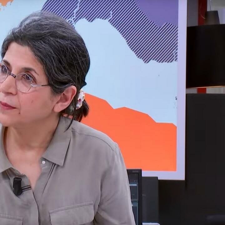 این تصویر فردی را نشان می‌دهد که در یک محیط خبری یا استودیو قرار دارد. او دارای موهای خاکستری و آرایش ساده است و عینک دارد. لباس او رسمی و به رنگ ملایم است. پس‌زمینه تصویر نقشه‌ای از جهان یا بخش‌هایی از آن را نمایش می‌دهد که به نظر می‌رسد به موضوع خبر مربوط می‌شود. چهره این فرد نشان‌دهنده توجه و تمرکز است.