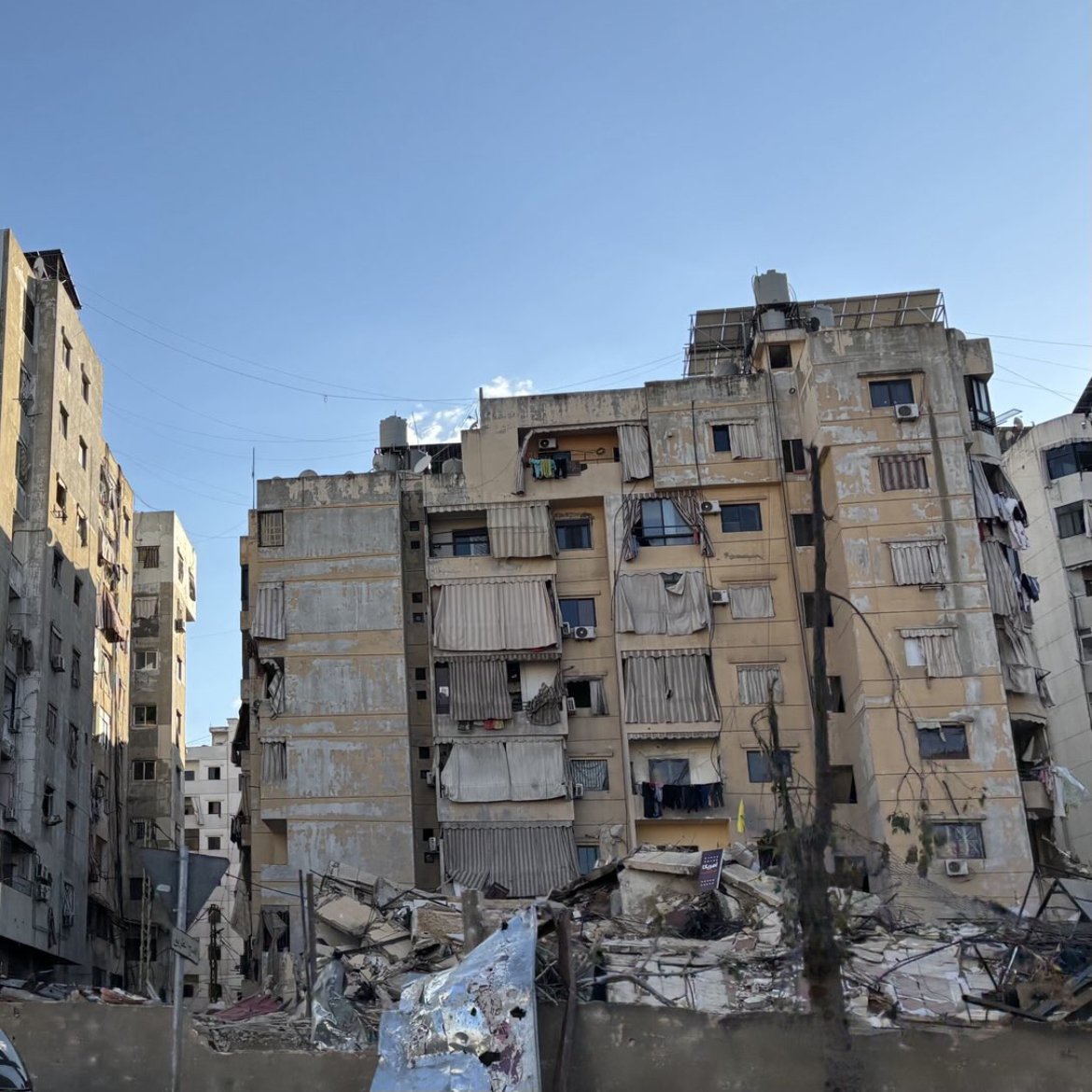 در این تصویر، یک ساختمان بزرگ با تصاویری از فردی که احتمالاً یک شخصیت عمومی است، دیده می‌شود. در کنار آن، ساختمان‌های دیگری وجود دارند که به دلیل آسیب‌های ناشی از زلزله یا جنگ، خرابه و آسیب‌دیده به نظر می‌رسند. وضعیت بناها نشان‌دهنده تخریب و ویرانی قابل توجهی است. آسمان در حال حاضر روشن است و ابرهای سفیدی در آن دیده می‌شود. سبزینگی در اطراف نیز وجود دارد که ممکن است درختان یا گیاهان باشد.