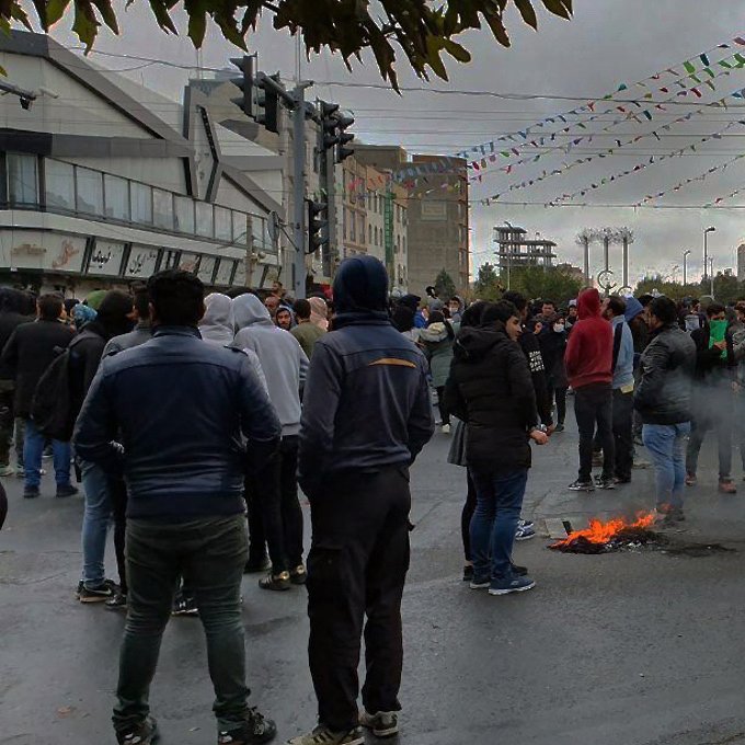 تصویر نشان‌دهنده یک تجمع اعتراضی در یک خیابان است. در پس‌زمینه، تعدادی از ساختمان‌ها دیده می‌شود و آسمان به رنگ خاکستری است. جمعیت زیادی از افراد در لباس‌های گرم در حال ایستادن یا حرکت کردن هستند. برخی از آنها ماسک به چهره دارند. در چند نقطه از خیابان، دود و شعله‌هایی به چشم می‌خورد که نشان‌دهنده وجود آتش است. همچنین، تزئینات و پرچم‌هایی در بالای خیابان دیده می‌شود که به نظر می‌رسد نشان‌دهنده یک مناسبت خاص یا جشنواره است. جمعیت به نظر می‌رسد که در حال تظاهرات یا اعتراض هستند.