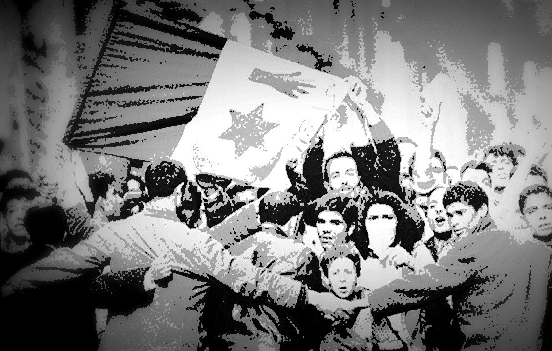 The image appears to depict a group of people gathered in a protest or demonstration. They are holding a flag that features a star and what looks like a hand symbol. The scene conveys strong emotions, with many individuals expressing solidarity and unity, possibly in a moment of significant political or social change. The image seems to be in black and white, which adds a sense of historical depth to the moment being captured.