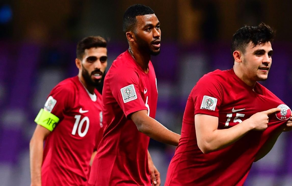 The image captures a moment from a football (soccer) match featuring players in red jerseys representing Qatar. One player, wearing the number 15, is celebrating and pulling his jersey up, possibly revealing a symbol or crest underneath. The player in the foreground seems excited and is likely celebrating a goal. Behind him, two other teammates are visible; one is wearing the number 10 and the other appears to be the team captain, as indicated by the captain's armband on his arm. The stadium setting suggests a significant match, likely during a tournament.