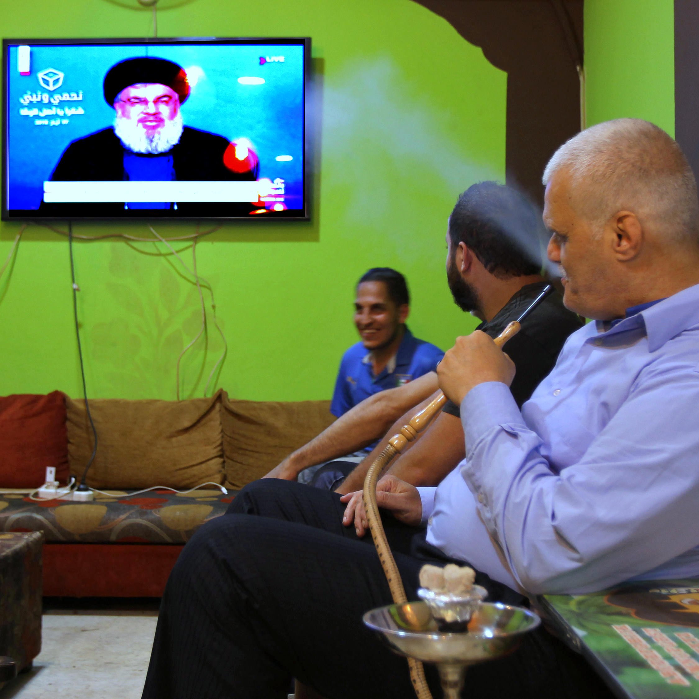 تظهر في الصورة مجموعة من الأشخاص يجلسون في غرفة معيشة مريحة، حيث يجلس رجلٌ على الأريكة وهو يدخن الشيشة. خلفه، يظهر تلفاز يُبث عليه صورة لشخص معروف يتحدث. الجدران تؤدي إلى أجواء دافئة، بينما الركن الآخر يُظهر شخصًا ضاحكًا. الألوان في المكان تتنوع بين الأخضر والبني، مما يعطي شعورًا بالراحة.