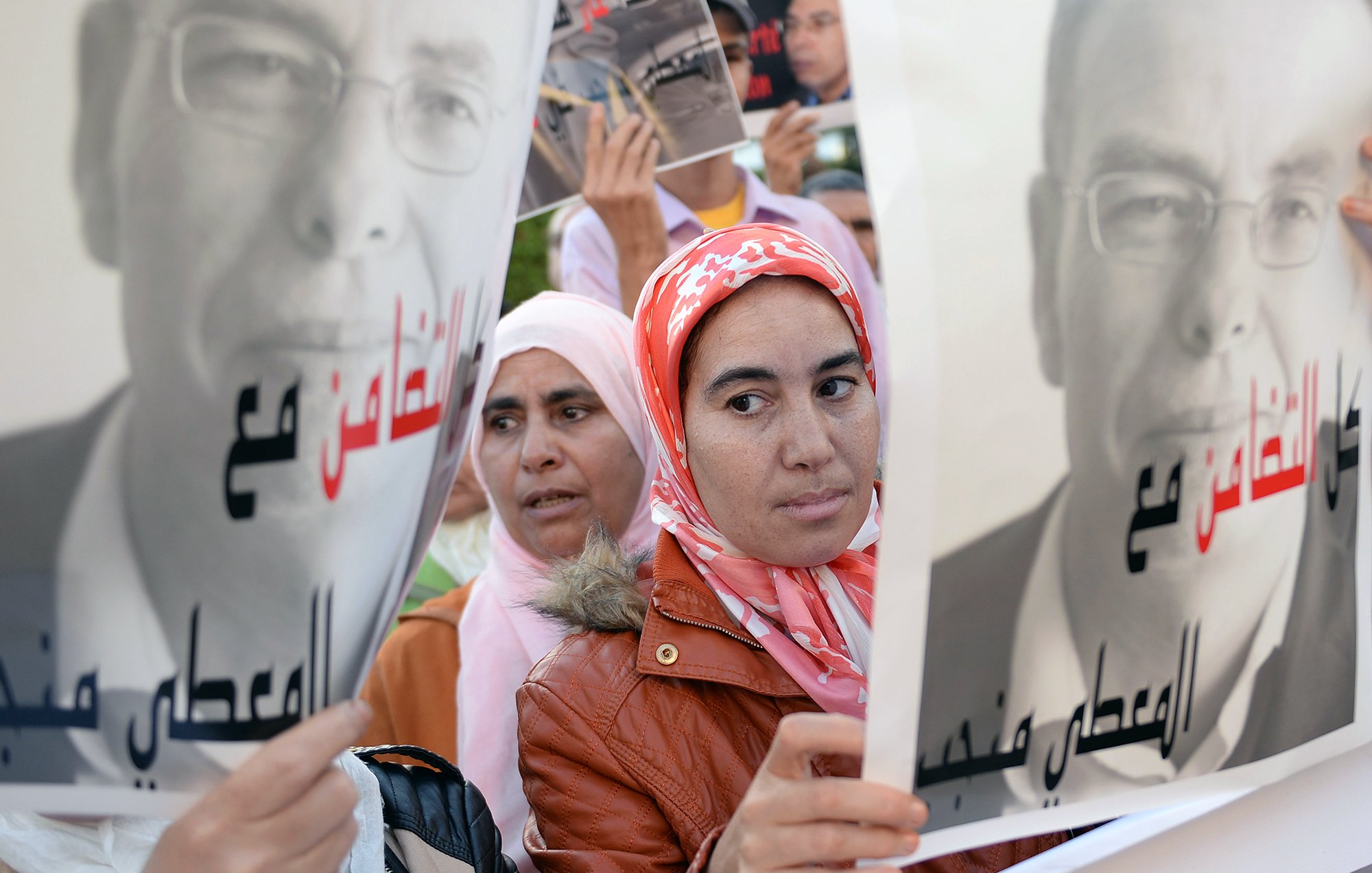 L'image présente des manifestants tenant des pancartes. Au premier plan, une femme avec un foulard rose regarde l'objectif, tandis qu'une autre femme, en arrière-plan, semble également attentive. Les pancartes affichent des portraits en noir et blanc d'un homme, accompagnés de slogans en arabe. L'ambiance suggère un rassemblement ou une manifestation, probablement pour défendre une cause ou exprimer une opinion.