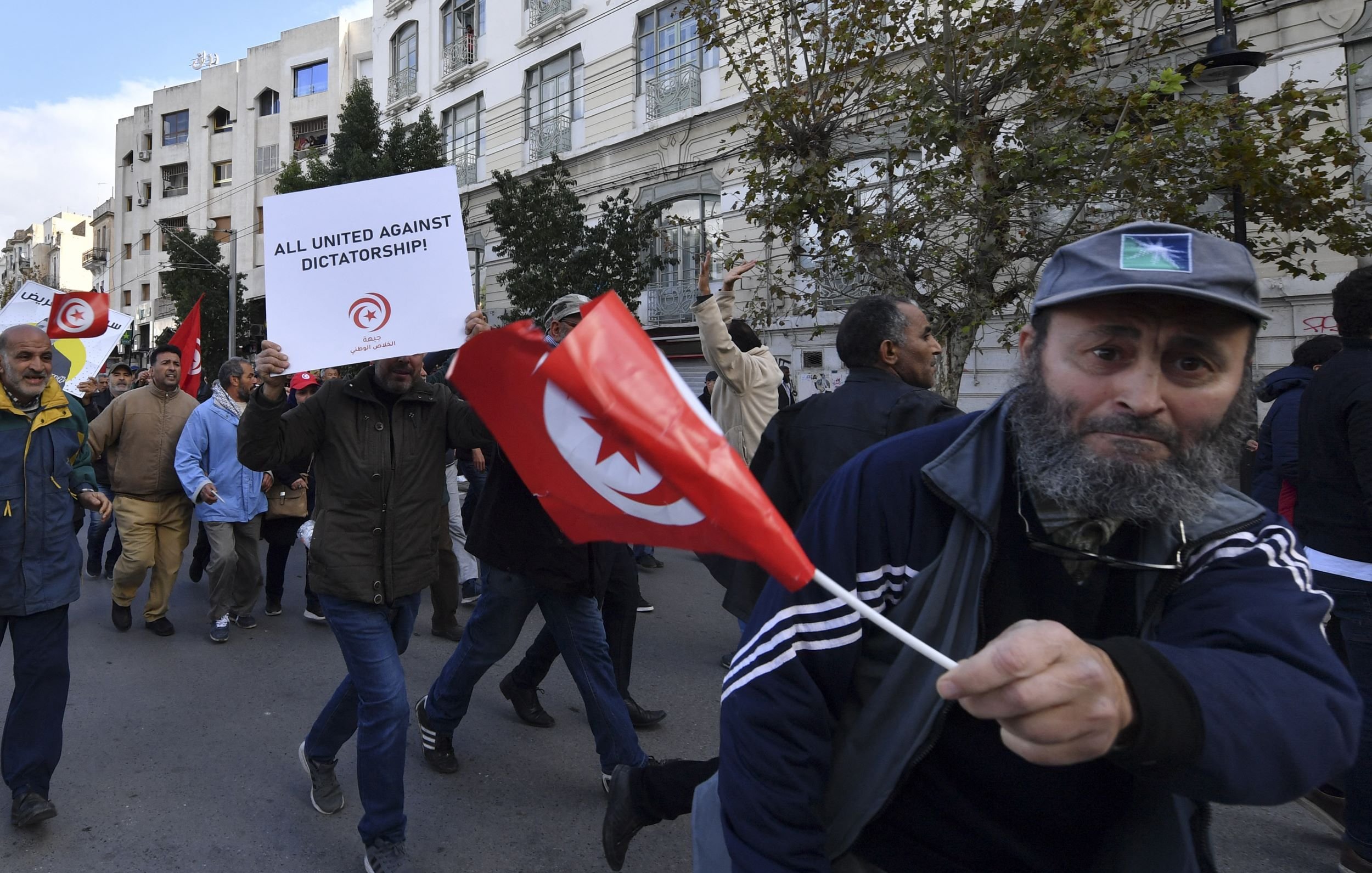 The image depicts a protest scene, likely in Tunisia. A crowd of people is marching down a street. Some participants are holding signs and flags. One prominent sign reads "ALL UNITED AGAINST DICTATORSHIP!" indicating the protest's focus on political issues. Among the crowd, a man stands out, waving a Tunisian flag and wearing a baseball cap and a jacket. The overall atmosphere suggests a movement against authoritarian rule, with participants expressing their collective dissent.