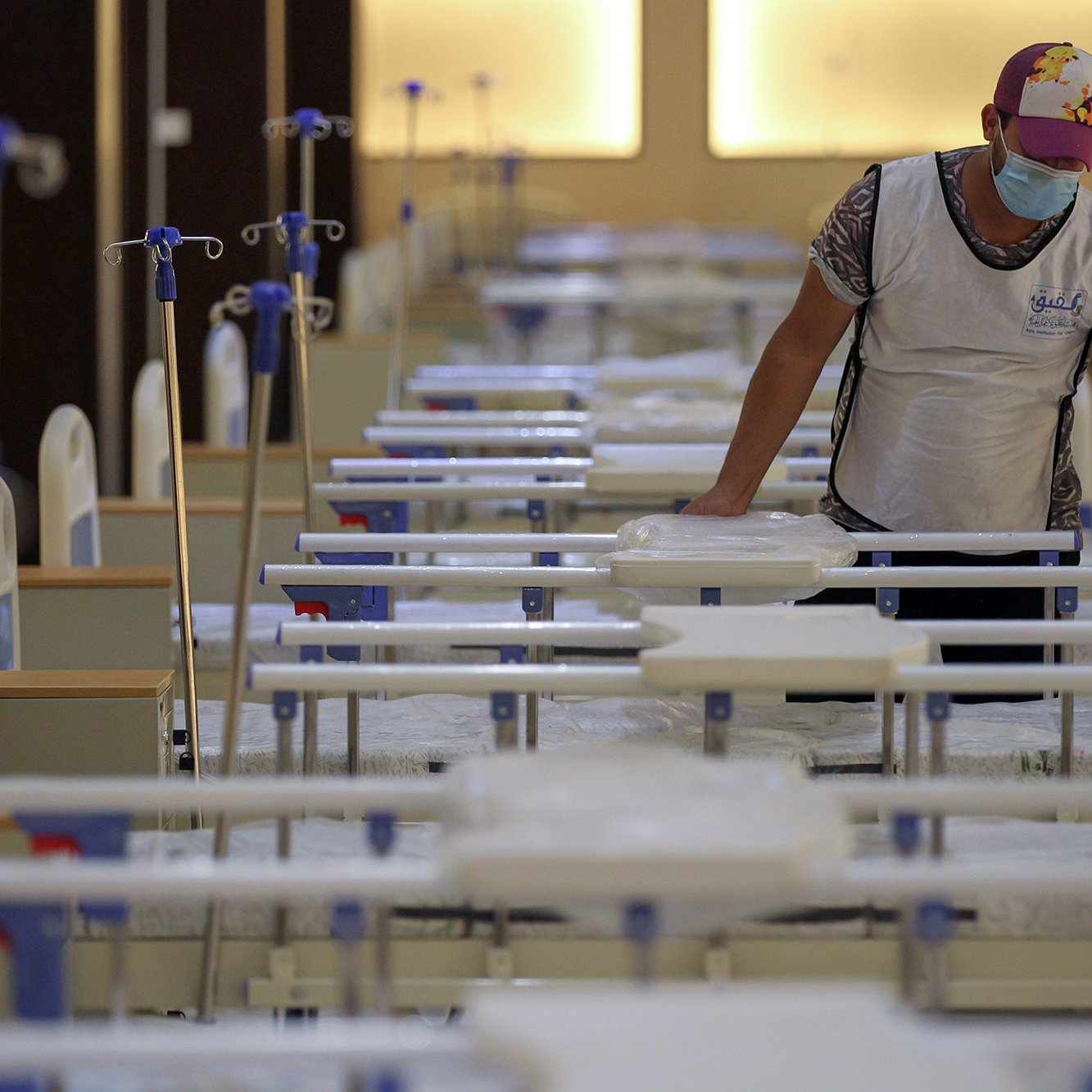 La imagen muestra a un hombre que está de pie junto a una serie de camas dispuestas en una sala. El hombre lleva una mascarilla y un chaleco, mientras que las camas están organizadas en filas, con sábanas y equipos médicos visibles. La iluminación es suave, lo que sugiere un ambiente de cuidado hospitalario. La escena da la impresión de un espacio preparado para atender pacientes.