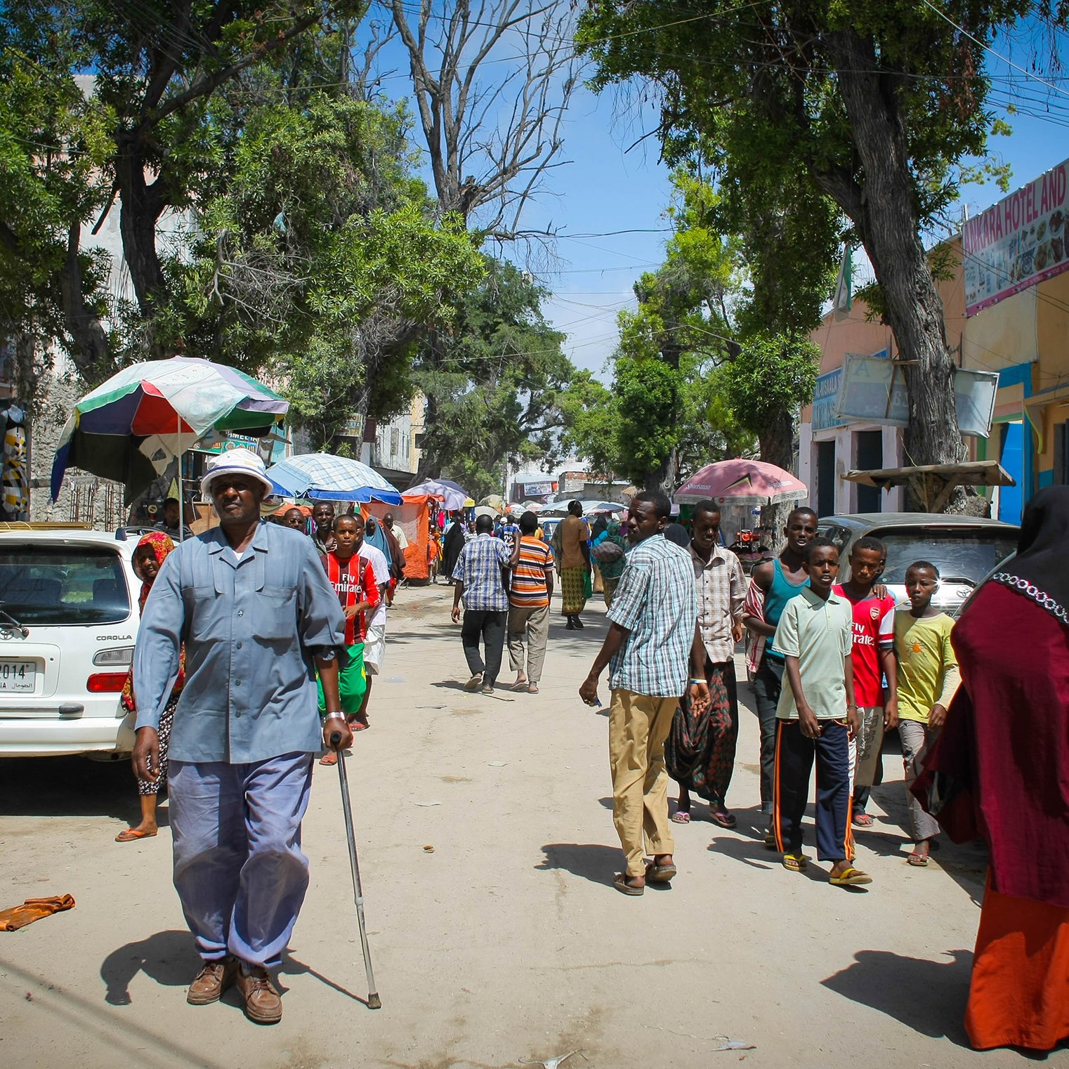 L'image montre une rue animée avec des groupes de personnes marchant. On peut voir un homme âgé utilisant une canne, habillé en bleu, qui semble avancer lentement. Plusieurs personnes sont dispersées le long de la rue, certaines se trouvent près de voitures garées. Des ombrelles colorées sont visibles, probablement pour se protéger du soleil. Les bâtiments en arrière-plan semblent être de petits commerces ou habitations. L'ambiance générale dénote une vie quotidienne dynamique dans un environnement urbain.