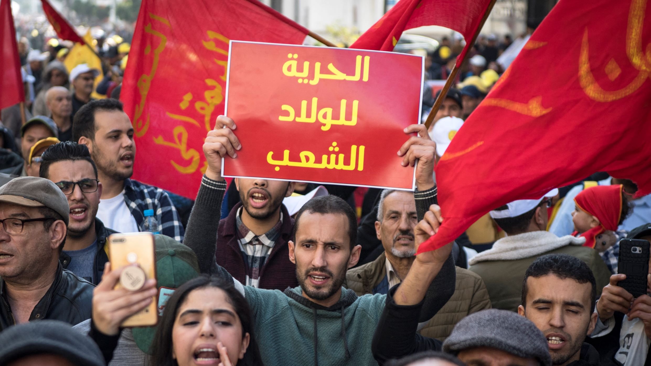 Nell'immagine si vede una manifestazione di protesta affollata, con persone che sventolano bandiere rosse. In primo piano, un giovane tiene in alto un cartello con scritto in arabo "libertà per il popolo". Altre persone intorno a lui sembrano partecipare attivamente, alcune con telefoni in mano per immortalare il momento. L'atmosfera è carica di emozione e determinazione, riflettendo il desiderio di cambiamento e di diritti.