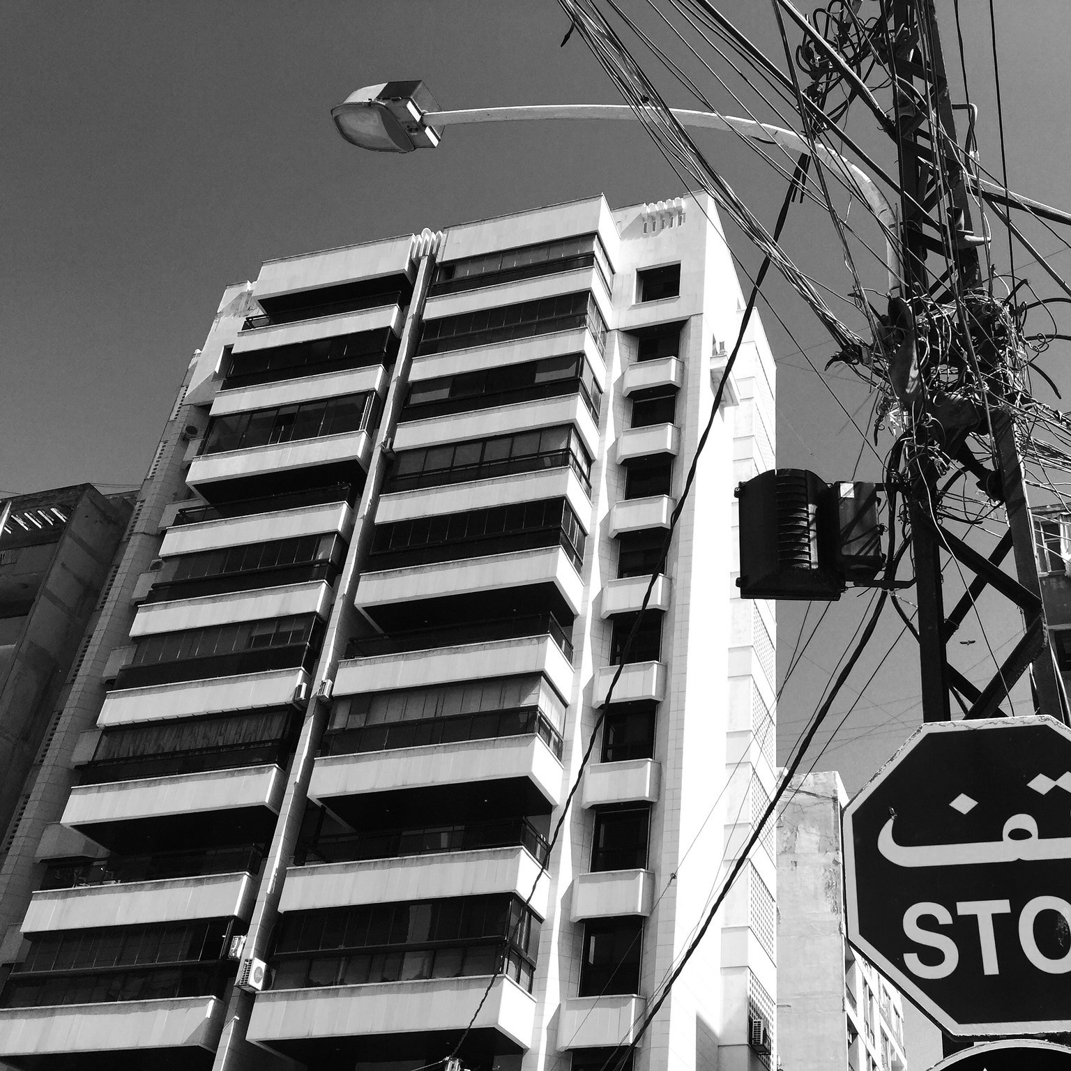 این تصویر یک خیابان شهری را نشان می‌دهد که در آن یک تابلو ایست با نوشته "قف" (STOP) قرار دارد. در پس‌زمینه، یک ساختمان بلند و مدرن به همراه خطوط برق در آسمان دیده می‌شود. رنگ تصویر سیاه و سفید است که به آن حال و هوای خاصی می‌بخشد. ظاهر شهر و معماری ساختمان‌ها، نمایی از زندگی روزمره شهری را ارائه می‌دهد.