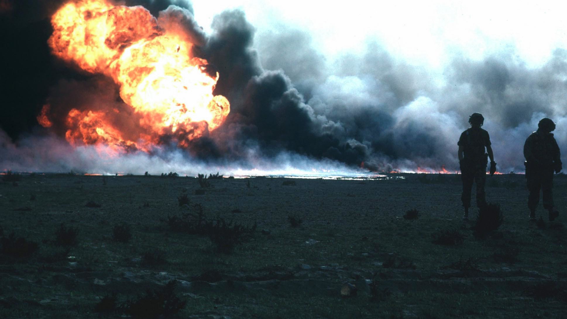 L'image montre une scène de destruction avec un grand incendie au loin. Des flammes vives jaillissent dans un nuage de fumée noirâtre. Au premier plan, deux silhouettes de soldats sont visibles, marchant côte à côte. Le paysage semble aride, avec peu de végétation. L'atmosphère est sombre et dramatique, soulignant l'impact de l'explosion.