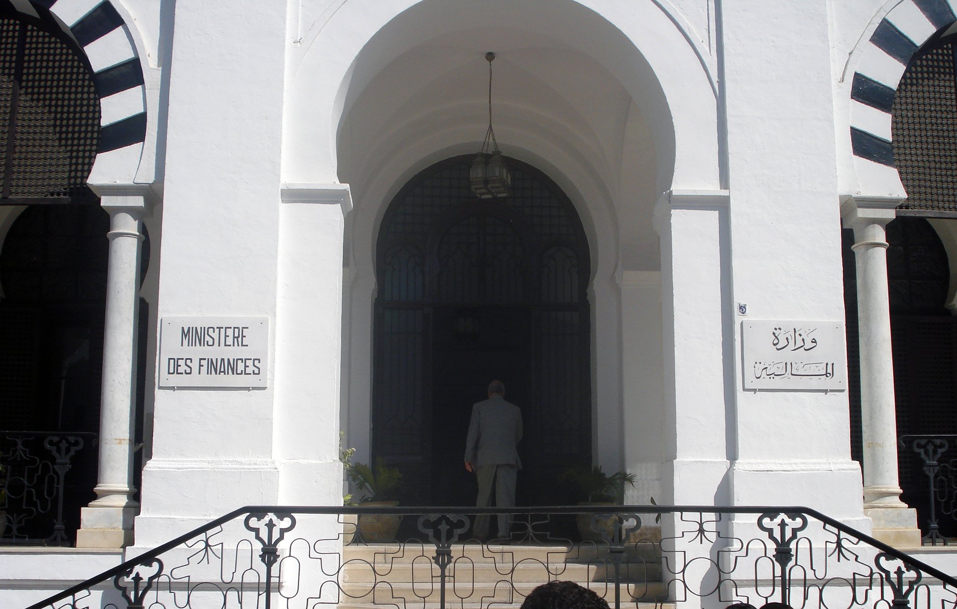 L'image montre l'entrée d'un bâtiment qui semble être le ministère des Finances, avec des inscriptions en français et en arabe. Le bâtiment est de style architectural classique, caractérisé par des murs blancs et des éléments décoratifs. On peut voir une grande porte d'entrée, entourée de fenêtres avec des grilles artistiques. À l'avant, une personne est visible, tournant le dos, s'approchant de l'entrée. Des plantes ornent l'espace autour des marches qui mènent à la porte.