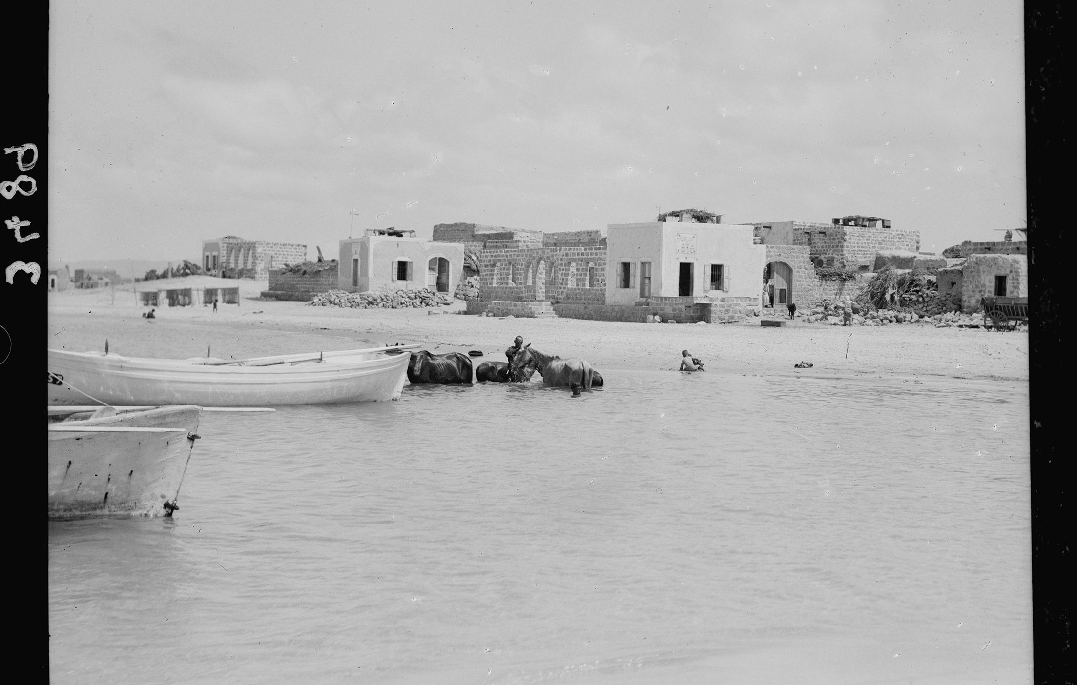 تظهر في الصورة مشهداً ساحلياً يضم مجموعة من المراكب الصغيرة الراسية على المياه. في الخلفية، توجد مبانٍ قديمة إلى جانب بعض الهياكل المتبقية، ما يدل على وجود مجتمع ساحلي سابق. يُرى بعض الأشخاص، بما في ذلك شخص يركب على ظهر حيوان، مما يعطي انطباعًا عن الحياة اليومية في هذا المكان. تتسم السماء بالغيوم المتفرقة، مما يوحي بأن الصورة ربما تم التقاطها في يوم مشمس.