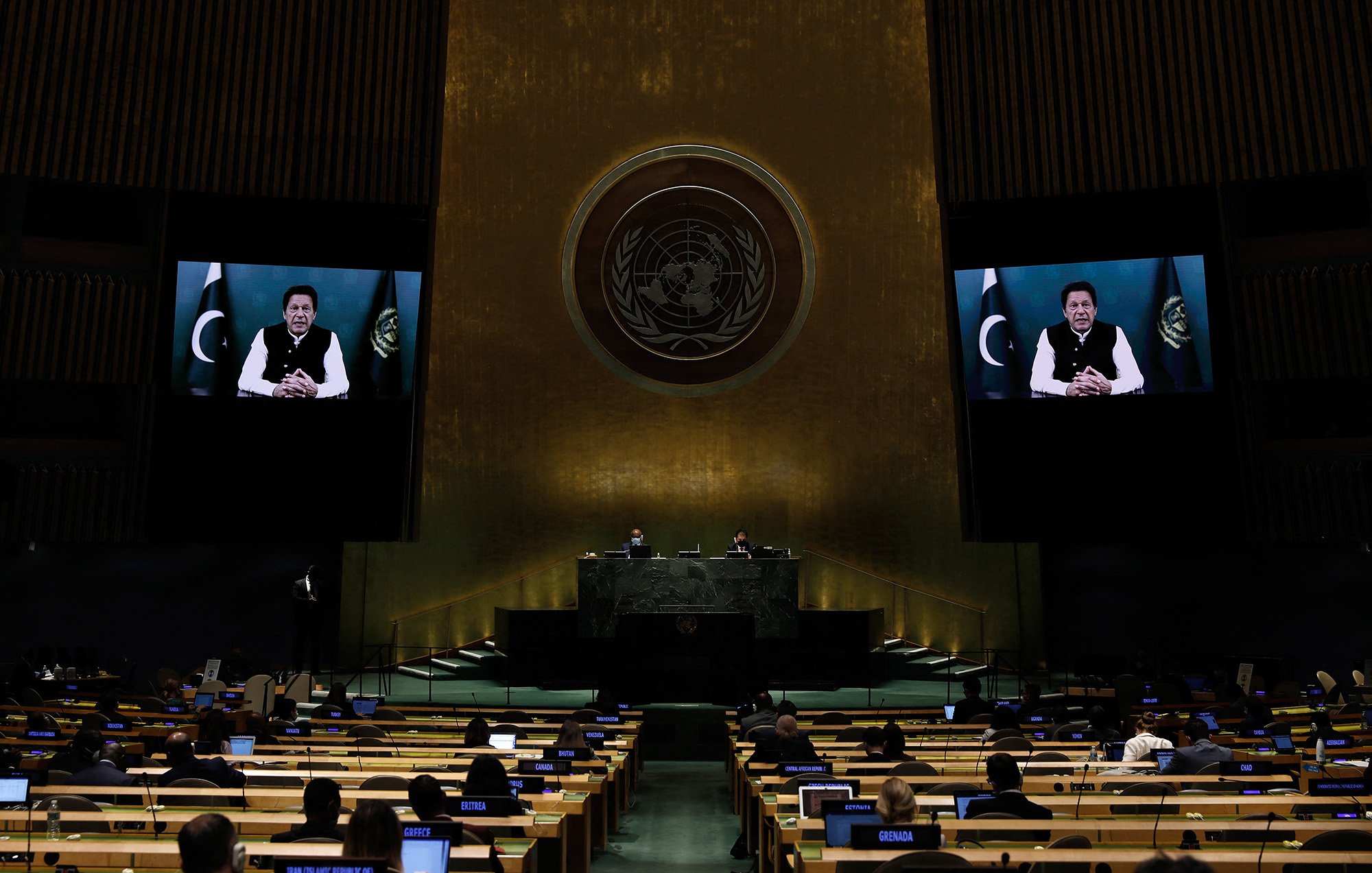 The image depicts a scene from a United Nations assembly. In the foreground, there are delegates seated at rows of desks, facing the main podium. The backdrop features a large screen displaying a speaker, who appears to be addressing the assembly. The setting is characterized by the assembly hall's distinctive architecture, featuring high walls and the emblem of the United Nations. The atmosphere is formal and legislative, reflecting an important discussion or speech taking place.