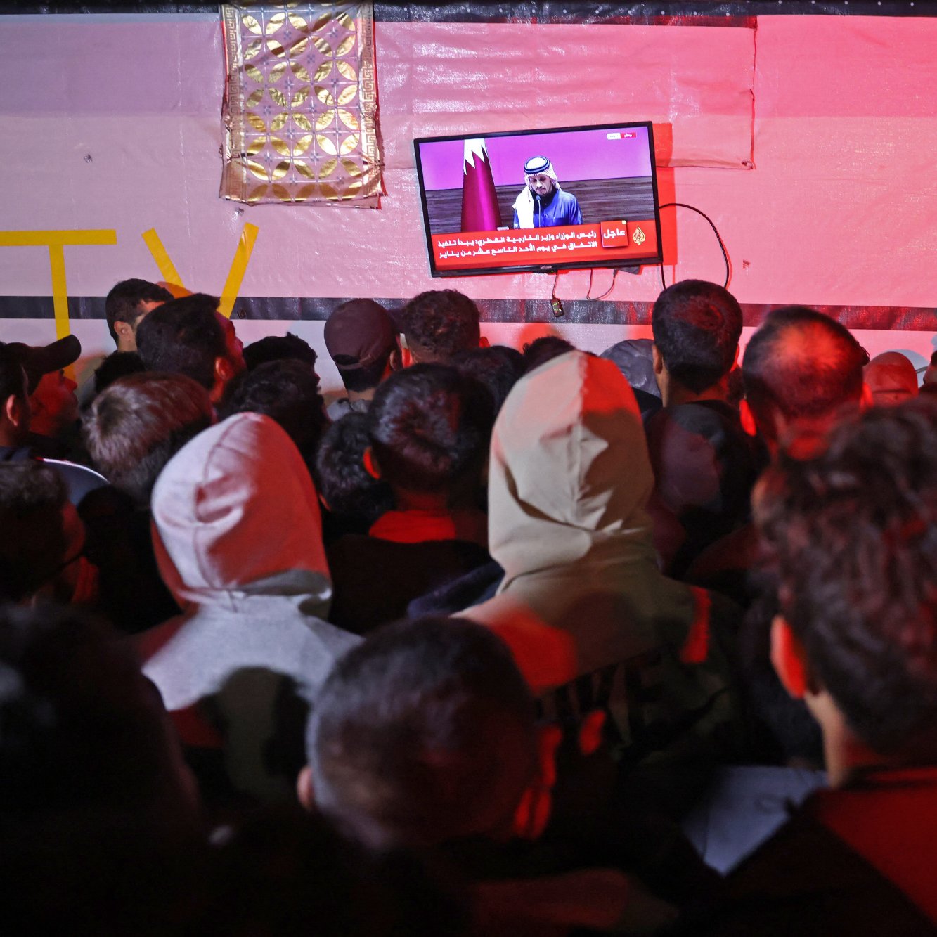 در تصویر، گروهی از افراد در حال تماشای تلویزیونی هستند که بر روی دیواری نصب شده است. تلویزیون یک برنامه خبری را نمایش می‌دهد و یک سخنران در حال صحبت است. افراد به سمت صفحه تلویزیون خیره شده‌اند و جو عمومی تصویر نشان‌دهنده توجه و کنجکاوی آن‌هاست. فضای اطراف کمی تاریک و نور محیطی کم است، به طوری که نور تلویزیون بیشتر مشخص است. برخی از افراد در لباس‌های گرم، نظیر هودی و کلاه، حضور دارند. یکی از افراد در سمت راست با دوربین در حال عکاسی است.