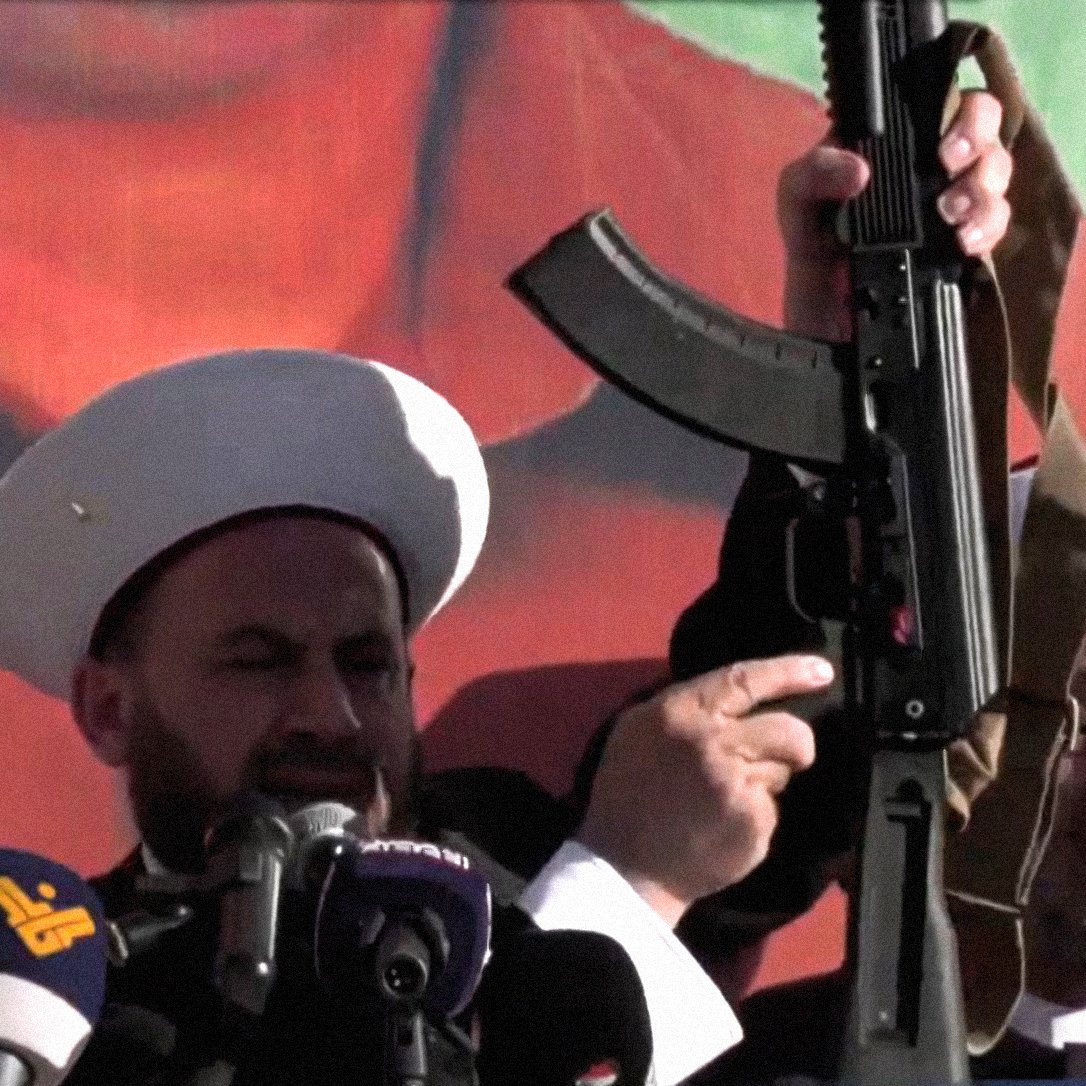 L'immagine mostra due uomini in abiti tradizionali, entrambi con copricapi bianchi, che sembrano partecipare a un evento pubblico o a una manifestazione. Uno di loro tiene in mano un fucile d'assalto, mentre l'altro osserva. Sullo sfondo c'è un grande murale o una bandiera che potrebbe rappresentare simboli politici o culturali. L'atmosfera sembra intensa e carica di emozione, con un focus sulla figura che tiene l'arma.