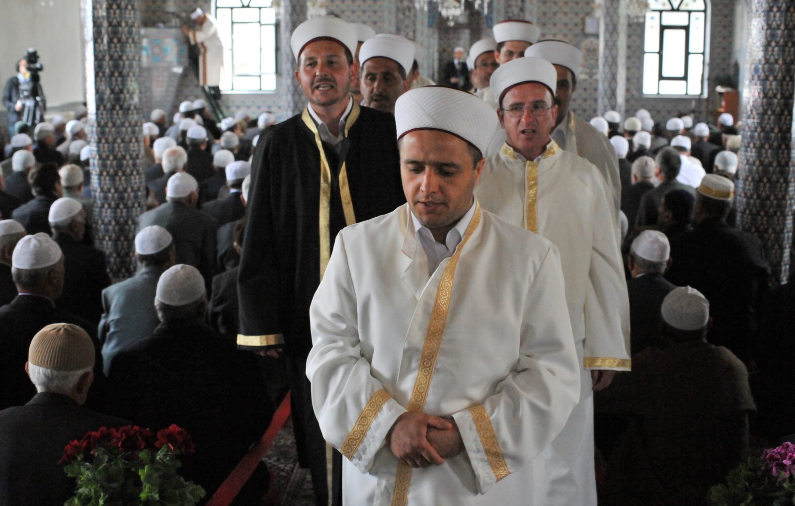 L'image montre un groupe d'hommes portant des vêtements traditionnels, notamment des robes blanches et des turbans. Ils semblent être en procession, probablement lors d'un événement religieux. En arrière-plan, de nombreuses personnes sont assises, probablement en prière ou en attente d'une cérémonie. L'architecture de la pièce inclut des motifs décoratifs et des fenêtres qui laissent entrer la lumière, créant une atmosphère solennelle.