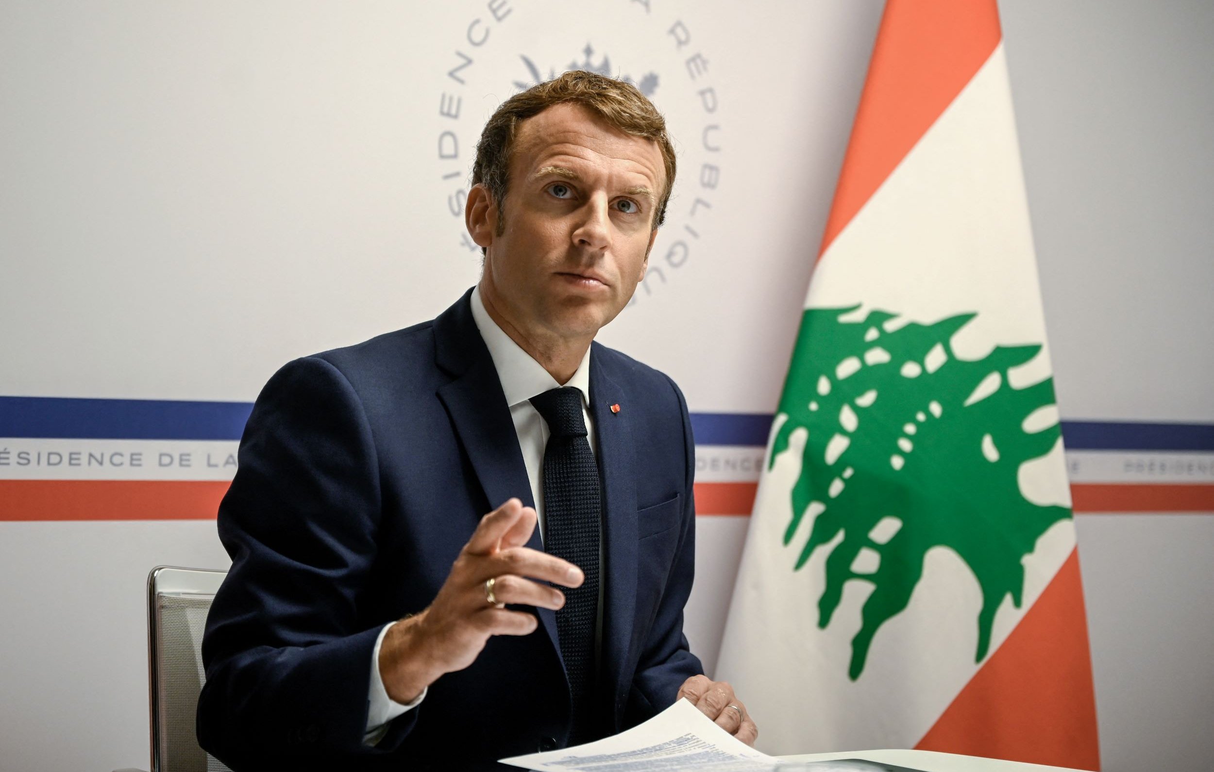 در تصویر، یک مرد با موی تیره و لباس رسمی (کت و شلوار) دیده می‌شود که به سمت دوربین نگاه می‌کند. او دارای ظاهری جدی و متفکر است و با دستش در حال بیان نقطه نظری است. در پس‌زمینه، پرچم لبنان و نشان رسمی نشان داده شده است. به نظر می‌رسد که این فرد در یک جلسه یا کنفرانس خبری مشغول سخنرانی است.