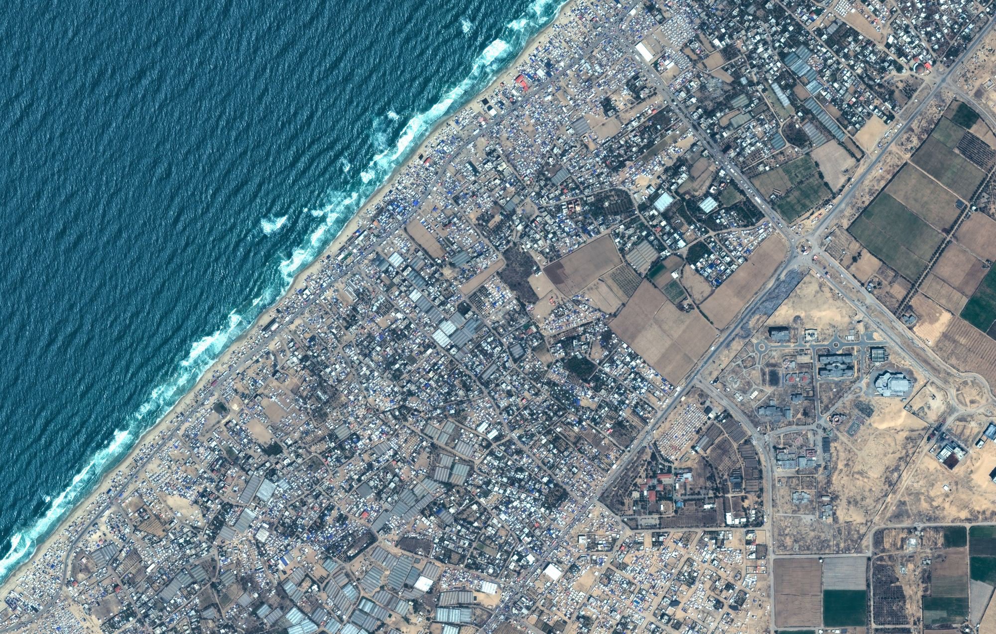 L'image montre une vue aérienne d'une zone côtière. En haut à gauche, on peut voir l'océan avec des vagues qui se brisent sur la plage. La partie inférieure de l'image présente des terrains variés, incluant des espaces urbanisés avec des routes et des bâtiments, ainsi que des zones agricoles avec des champs. La transition entre la zone bâtie et la nature est également visible. C'est une représentation d'un paysage où les zones urbaines rencontrent la côte.