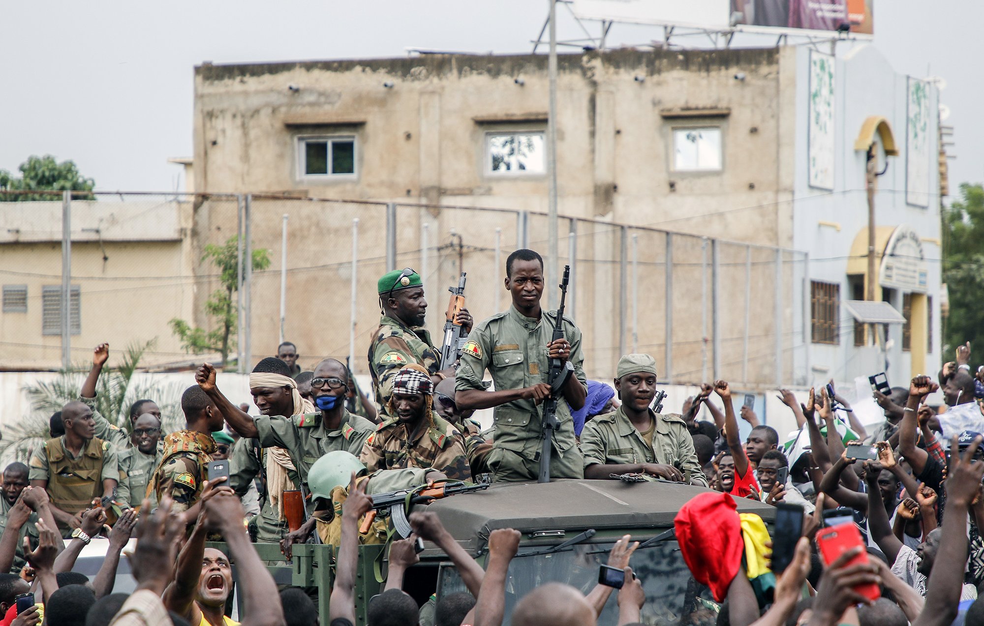 L'image montre un groupe de soldats, armés, sur un véhicule militaire, entourés par une foule enthousiaste. Les soldats semblent célébrer ou soutenir une cause, tandis que les membres de la foule lèvent les bras et expriment leur joie ou leur enthousiasme. En arrière-plan, on peut apercevoir des bâtiments et une atmosphère de mobilisation populaire.