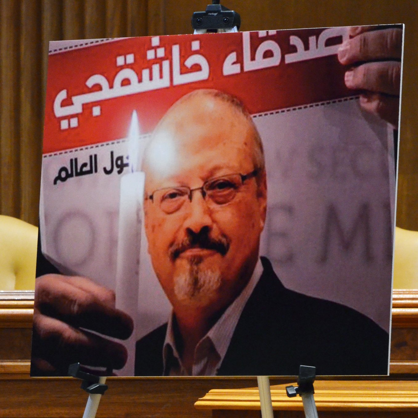 The image features a large poster of a man holding a candle, set against a background with text in Arabic. The poster is likely used in a context related to remembrance or tribute. The setting appears formal, possibly in a legislative or governmental chamber, indicated by the presence of chairs and the overall decor. The image emphasizes themes of justice and memory.