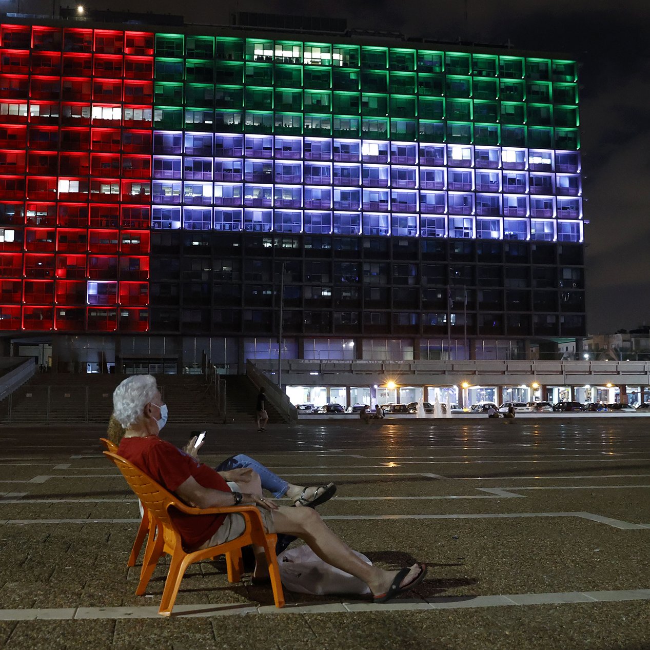 En la imagen se puede ver a un hombre mayor sentado en una silla de plástico anaranjada, mirando su teléfono móvil. Detrás de él, se destaca un gran edificio iluminado que muestra los colores de la bandera de México: rojo, blanco y verde. Es de noche, y el ambiente parece tranquilo con algunas personas caminando en el área. La arquitectura del edificio se aprecia moderna, y las luces crean un contraste vibrante con el cielo nocturno.