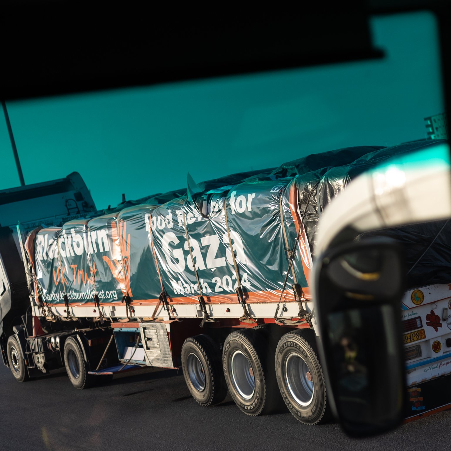 L'immagine mostra un camion lungo che trasporta una grande quantità di beni. Il veicolo è visibile da un angolo diverso, suggerendo che la foto potrebbe essere stata scattata da un altro veicolo. Sulla superficie del camion è presente un'informazione, probabilmente un messaggio di solidarietà o supporto, che menziona "Gaza" e "marzo 2024". Il contesto suggerisce un'operazione umanitaria o di aiuto. Sullo sfondo si possono vedere elementi di un paesaggio stradale.