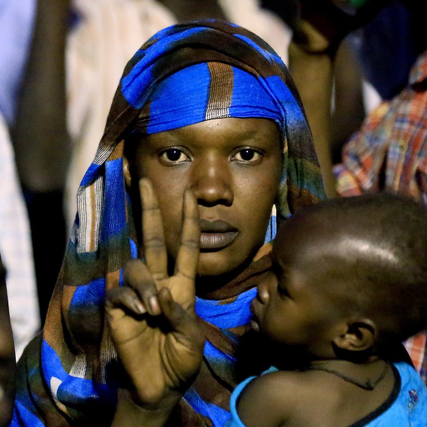 L'image montre une femme portant un foulard coloré, qui fait un signe de paix avec sa main. Elle tient un jeune enfant dans ses bras. L'arrière-plan semble flou, suggérant qu'elle se trouve dans une foule. Les expressions sur son visage et sa posture peuvent indiquer un moment de détermination ou de solidarité.