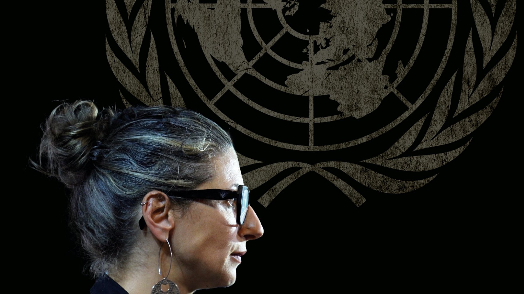 زنی با موهای خاکستری و عینک، در کنار نماد سازمان ملل، در حال صحبت کردن است.