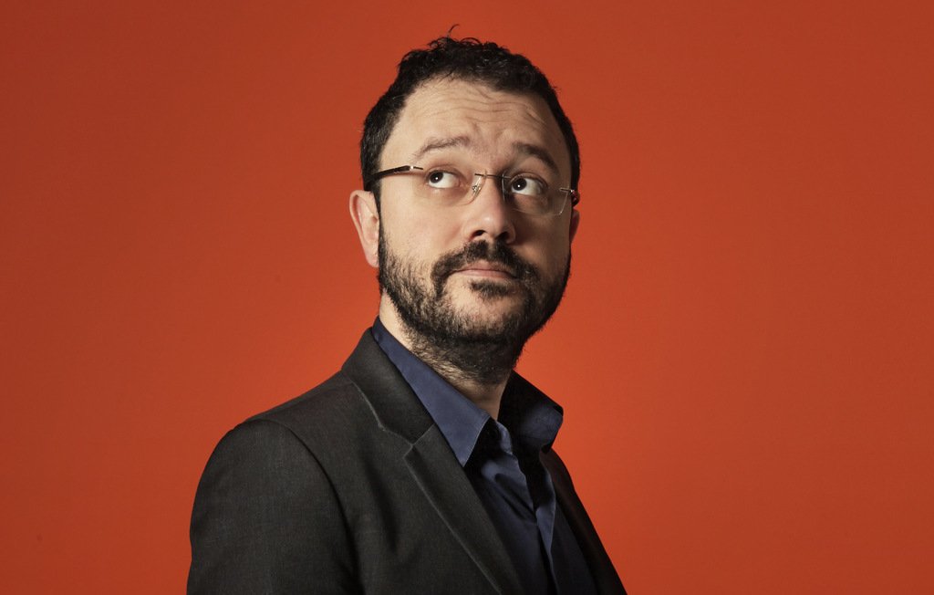 L'image montre un homme vêtu d'un costume noir sur une chemise bleu foncé, regardant légèrement sur le côté. Il a des lunettes, une barbe et un léger sourire. L'arrière-plan est orange, ce qui crée un contraste vif et dynamique avec sa tenue sombre. L'expression de son visage semble pensif ou intrigué.