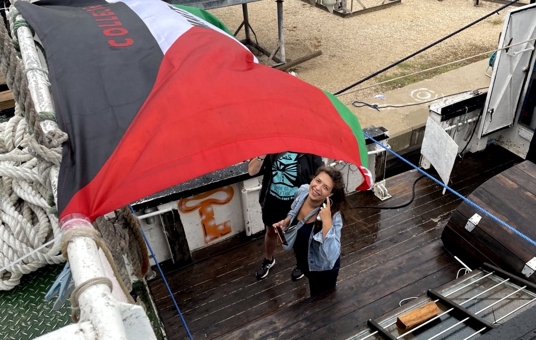 L'image montre deux personnes sur un bateau, sous un grand drapeau noir, rouge et vert. L'une des personnes sourit et pose pour la photo, tandis que l'autre semble tenir le drapeau. Le décor autour inclut du bois, des cordages et d'autres éléments d'un environnement nautique. L'atmosphère semble conviviale et dynamique.