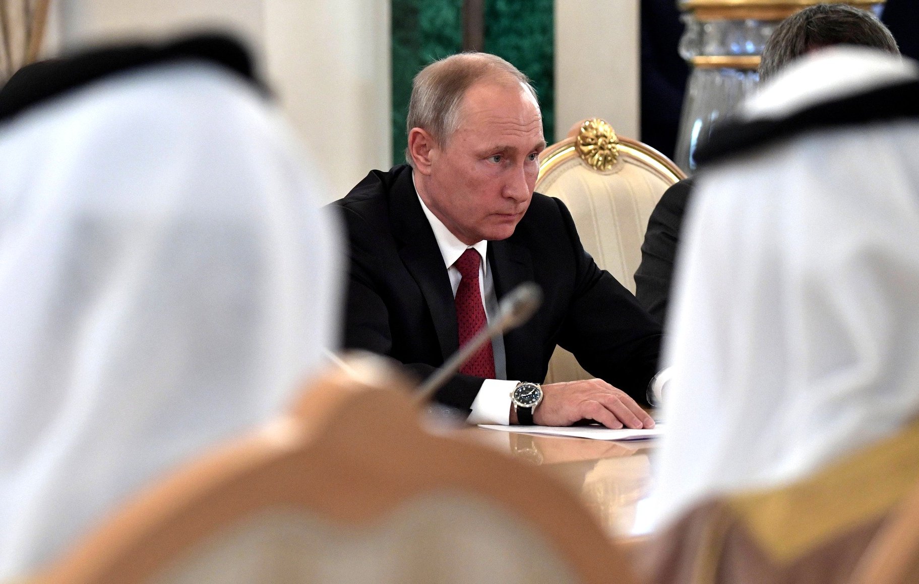 در این تصویر، فردی که به نظر می‌رسد ولادیمیر پوتین است، در حال نشستن در یک جلسه به سمت جلو نگاه می‌کند. او کت و شلوار مشکی به همراه یک رابعه قرمز به تن دارد. در اطراف او، چهره‌هایی از دیگر شرکت‌کنندگان در جلسه دیده می‌شود، اما جزئیات آن‌ها قابل تشخیص نیست. فضای عمومی جلسه جدی به نظر می‌رسد و میز مستطیلی به همراه دکوراتورهای رسمی دیده می‌شود.