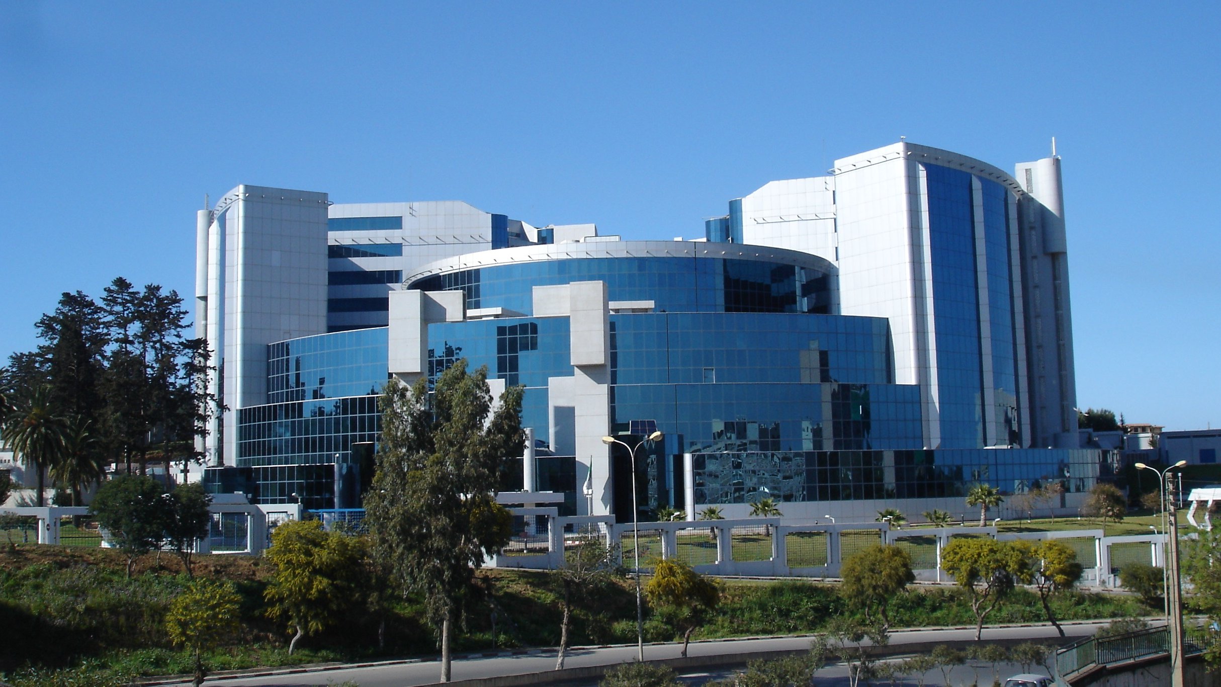 The image showcases a modern, multi-story building characterized by a sleek and reflective glass facade. The architecture features a combination of geometric shapes, with prominent curved and angular elements. Surrounding the building are landscaped areas with trees and greenery, contributing to an aesthetic appeal. The sky above is clear and blue, enhancing the overall vibrant look of the structure.