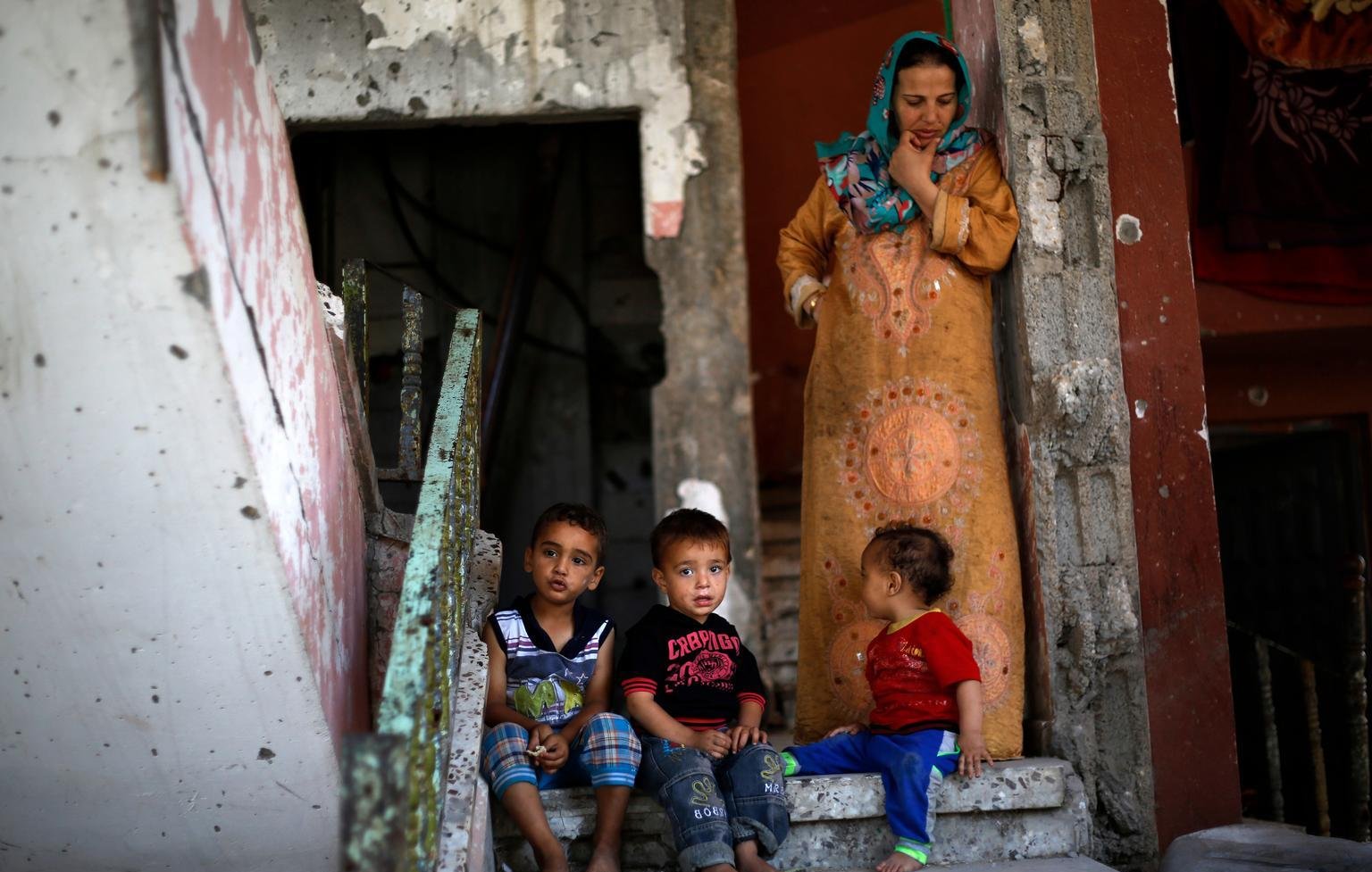 L'image montre une femme et plusieurs jeunes enfants assis sur des marches en béton. La femme est vêtue d'une robe longue, tandis que les enfants, assis près d'elle, portent des vêtements colorés. Le décor environnant semble ancien et en mauvais état, avec des murs écaillés et des escaliers. L'ambiance générale évoque une atmosphère de vie quotidienne dans un environnement modeste.