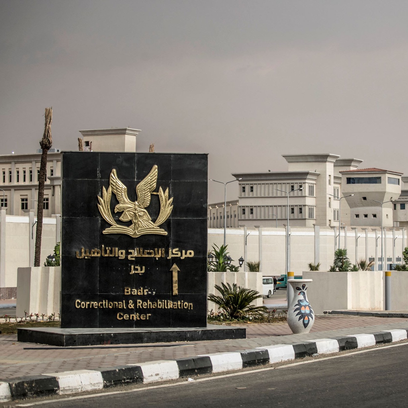 La imagen muestra un letrero grande y elegante que indica la entrada del "Centro de Corrección y Rehabilitación de Badr". El letrero tiene un diseño prominente con un símbolo de un águila en la parte superior. Al fondo se puede ver una construcción moderna y amplia, posiblemente el centro mismo, rodeada de muros altos y bien cuidados. La carretera está pavimentada y hay algunas decoraciones de cerámica a los lados. El cielo parece nublado, dando un ambiente de calma.