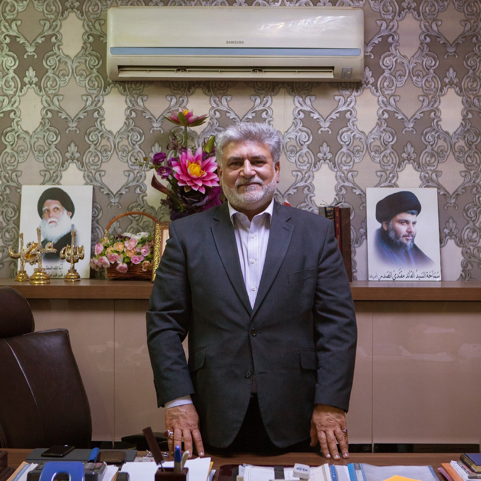 L'image montre un homme debout devant un bureau bien aménagé. Il porte un costume sombre et un sourire. Derrière lui, on peut voir des portraits de deux figures, ainsi qu'un arrangement floral. Le décor est élégant avec des murs ornés de motifs. L'ensemble dégage une atmosphère de professionnalisme et d'autorité.