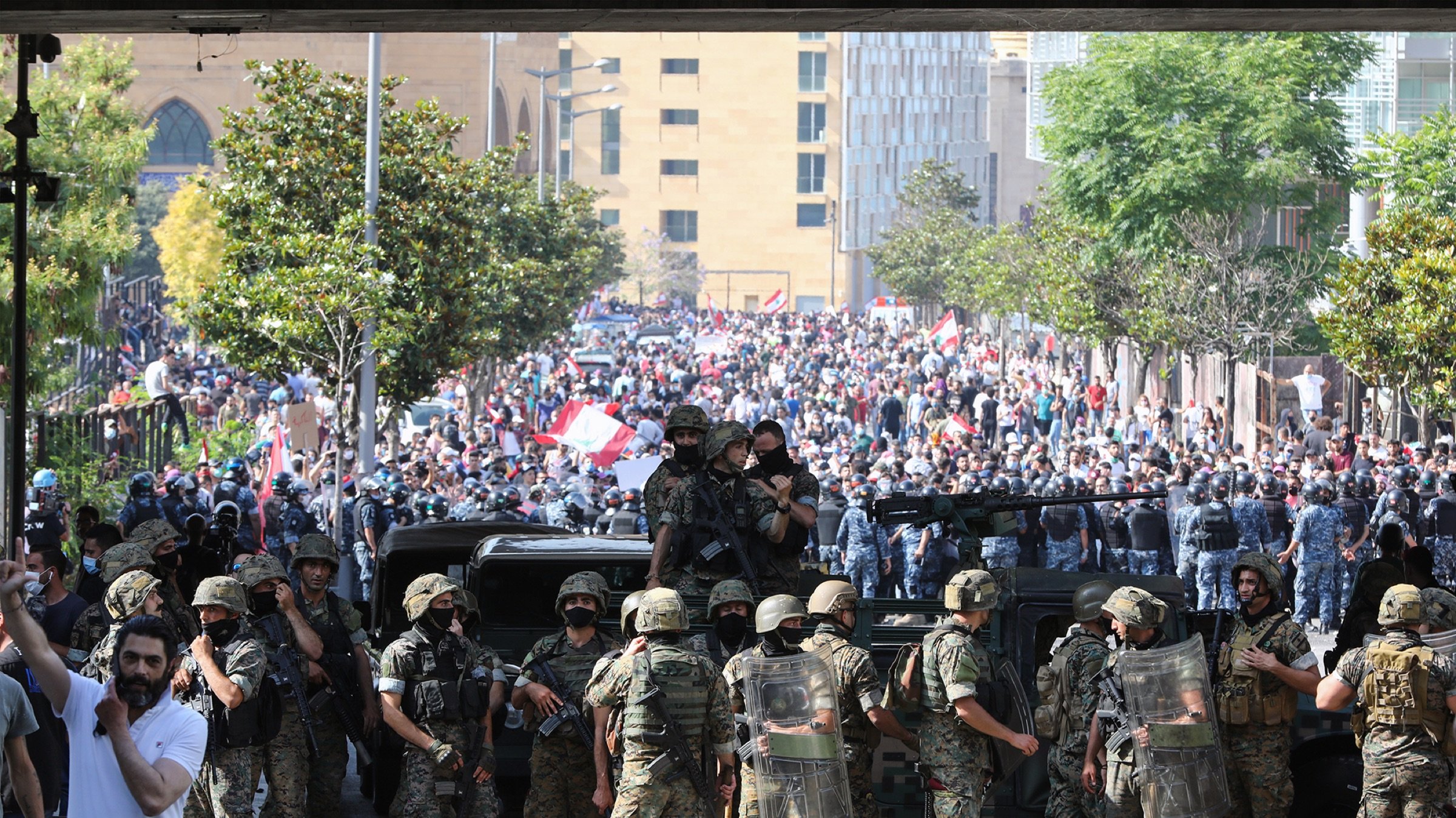 L'image montre une scène de protestation dans une ville, probablement à Beyrouth. Au premier plan, des soldats en uniforme sont alignés, portant des équipements de protection et des casques. Ils semblent faire face à une grande foule de manifestants qui se trouvent derrière eux. La foule est chargée d'énergie et porte des drapeaux, ce qui indique qu'il s'agit d'une manifestation pour une cause. Au fond, on peut voir des bâtiments urbains, ce qui ajoute un contexte à la scène. L'atmosphère est tendue, reflétant un moment significatif dans une dynamique sociale ou politique.